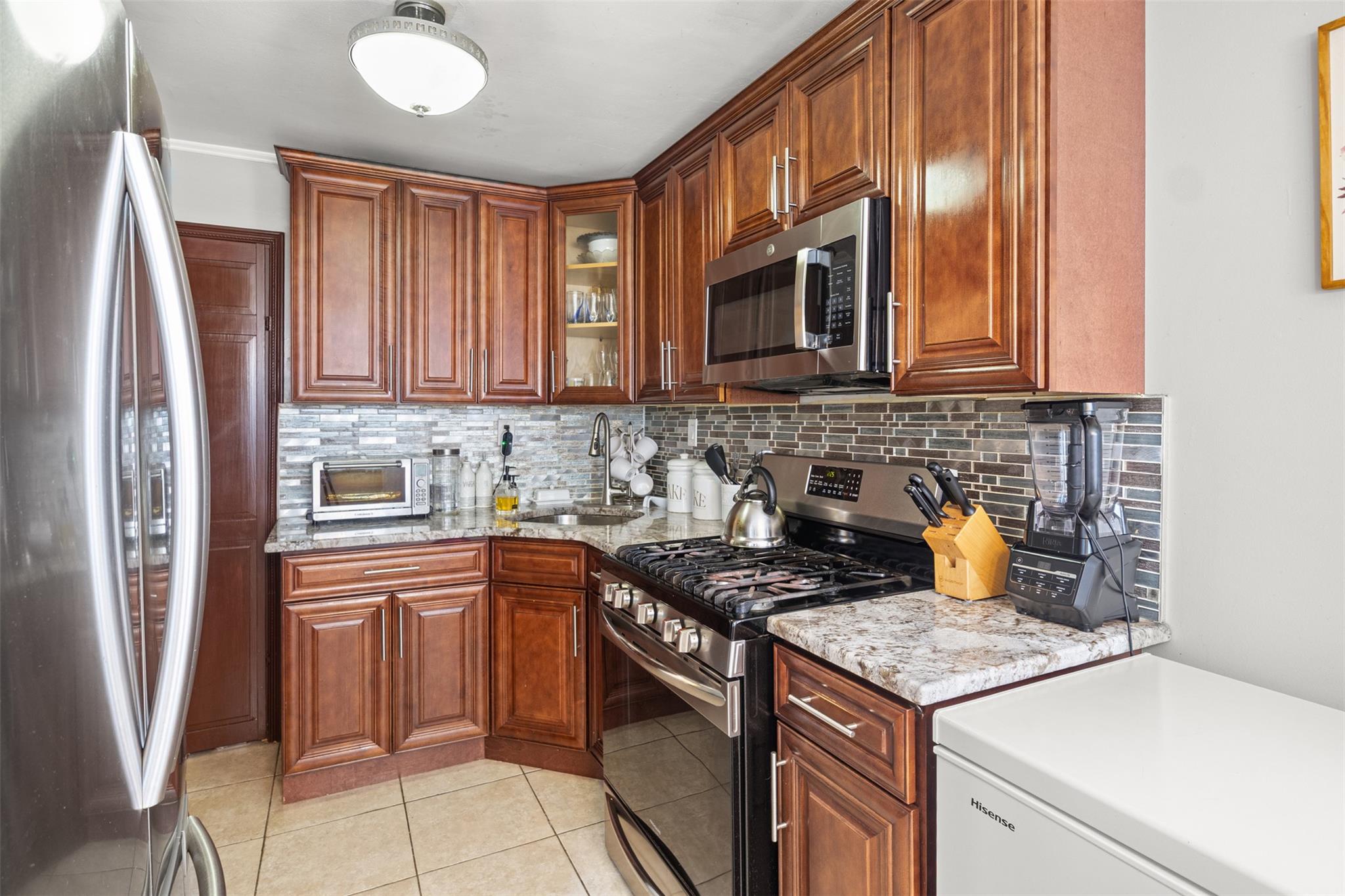 239-15 147th Road Queens, NY 11422 - Photo 5 of 28 Kitchen featuring appliances with stainless steel finishes, backsplash, light tile patterned flooring, glass insert cabinets, and light stone counters