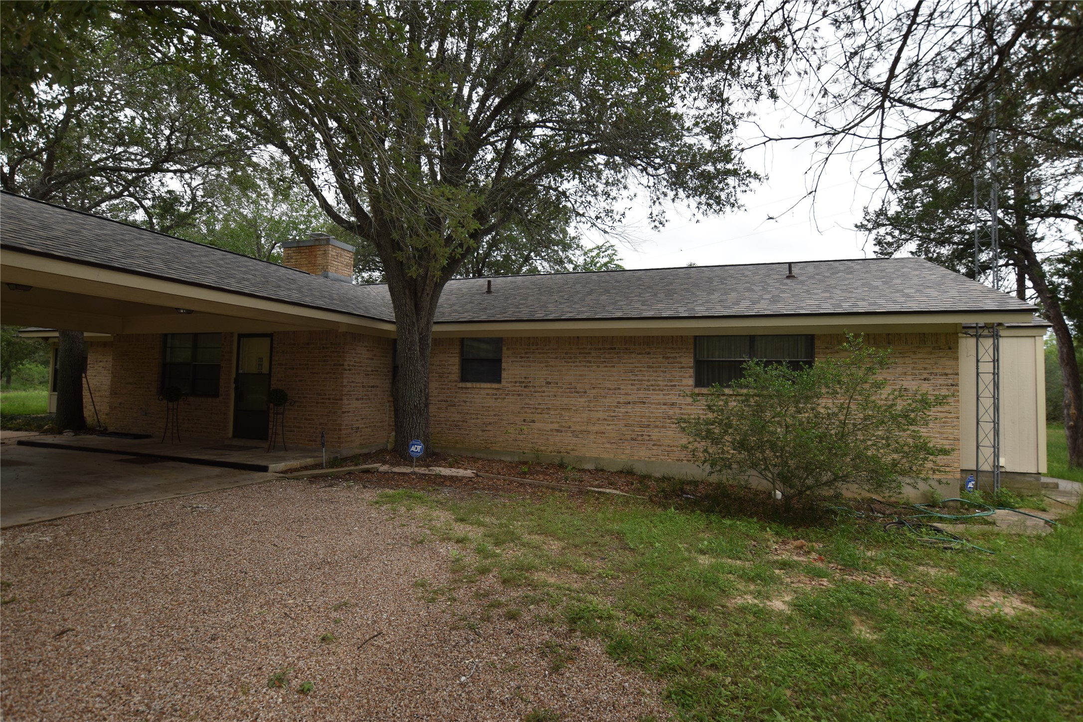 687 Fm 812 Road Red Rock, TX 78662 - Photo 3 of 40 Brick exterior featuring a covered carport, mature trees, a shingle roof, and a gravel driveway