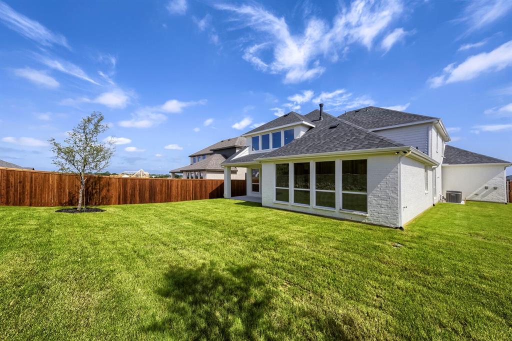 800 Blackberry Road Haslet, TX 76052 - Photo 26 of 28 Expansive green lawn bordering a tall privacy fence, featuring a young tree and a two-story residence with white brick and siding, a dark shingle roof, and multiple large windows