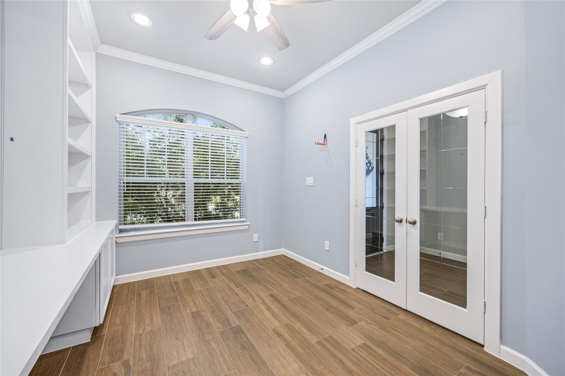 3604 Magnolia Crest Lane Spring, TX 77386 - Photo 7 of 50 Home office features a modern design with light blue walls, wood-look tile flooring, and built-in shelving. It has a large arched window with blinds for natural light and double glass doors, offering a bright and inviting space.