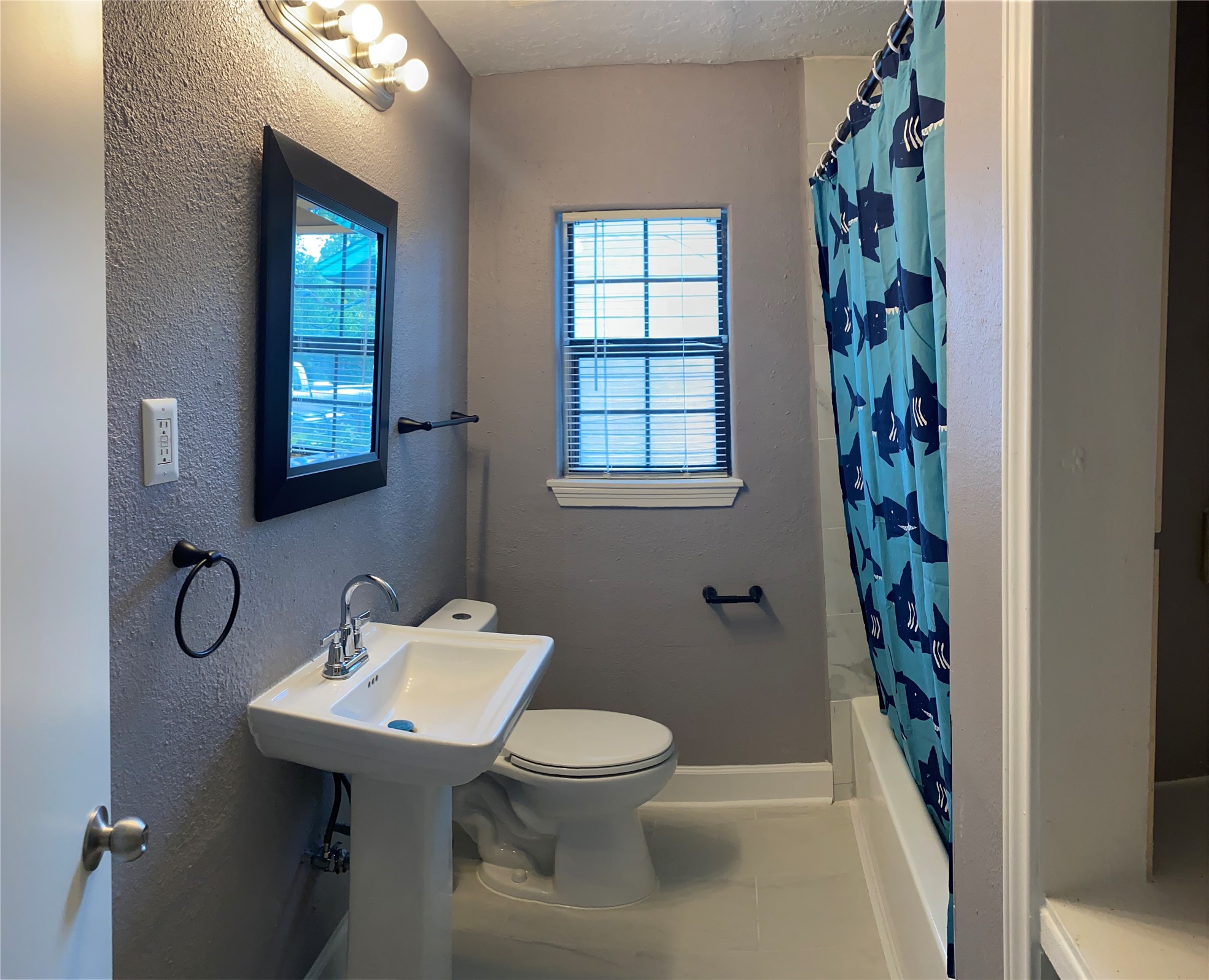 7547 Laura Koppe Road Houston, TX 77028 - Photo 9 of 11 This bathroom features a modern sink, a toilet, and a bathtub with a playful shark-themed shower curtain. It has a window for natural light and a dark-framed mirror, complemented by soft lighting.