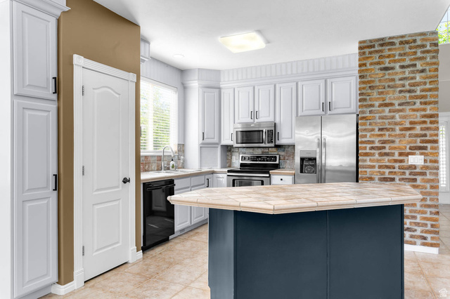 1417 Blacksmith Lane Draper, UT 84020 - Photo 15 of 57 Kitchen featuring a tiled island, white cabinetry, stainless steel appliances, and a brick-finish accent wall