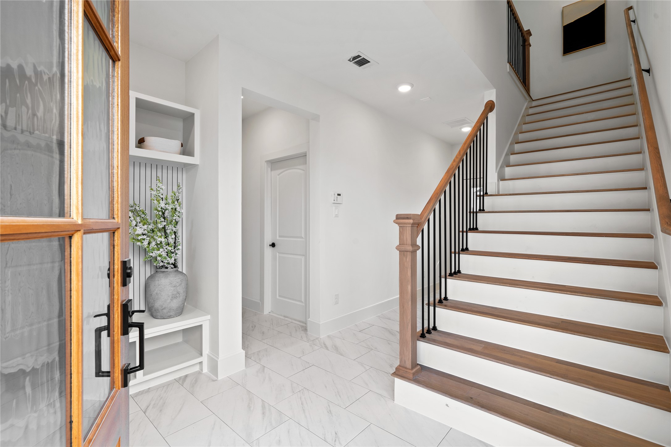 1614 Ojeman Road, Unit J Houston, TX 77055 - Photo 5 of 50 A bright entryway with a wooden front door, leading to a staircase with wooden steps and a metal railing. The space features white walls, tiled floors, and modern decor, creating a welcoming and contemporary atmosphere.