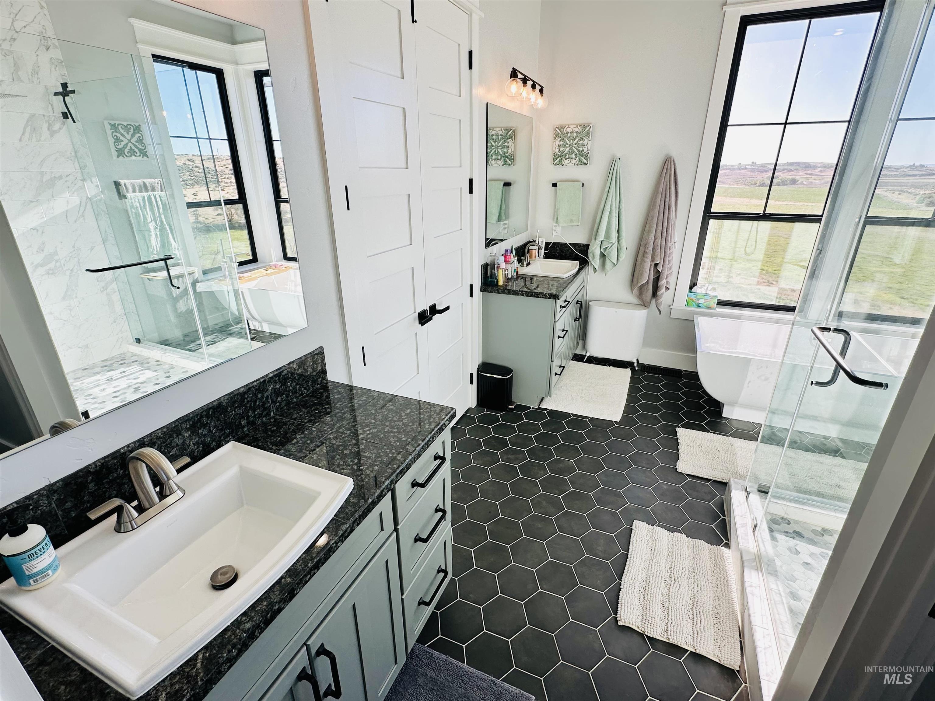 876 Coyote Gulch Road Adrian, OR 97901 - Photo 19 of 50 Bathroom featuring large hexagonal floor tiling, a freestanding soaking tub, a walk-in shower with clear glass enclosure, dual vanities with granite countertops, and multiple large windows