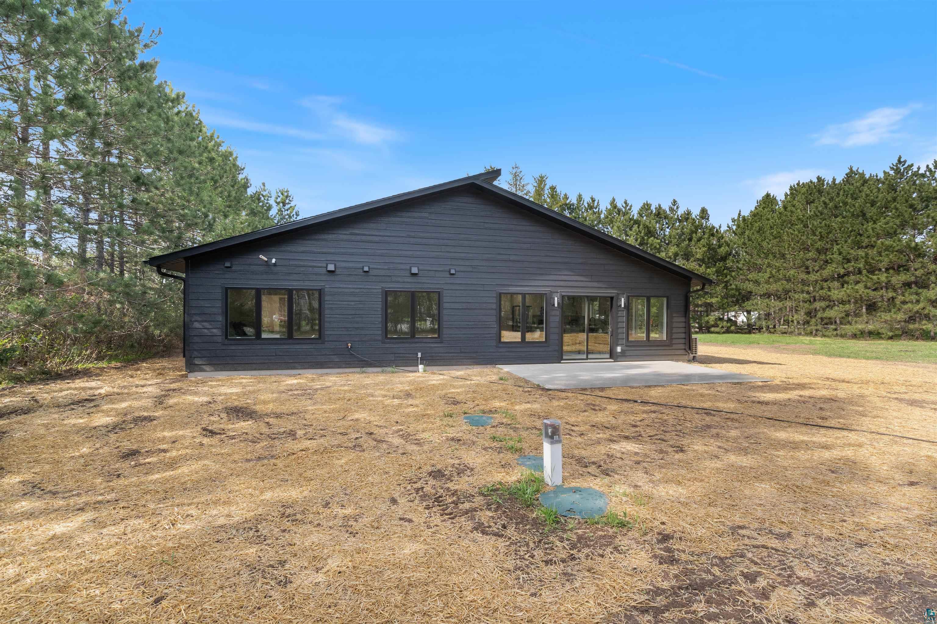 1386 Stephen Road Cloquet, MN 55720 - Photo 4 of 25 Modern black exterior with dark trim windows, a spacious concrete patio, a prominent sliding glass door, and extensive surrounding tree lines
