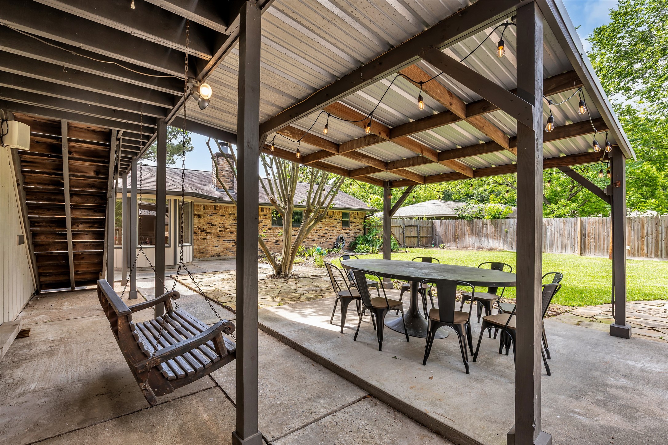 22502 Fields Lane Spring, TX 77389 - Photo 32 of 39 Outdoor patio with a covered pergola, featuring a spacious dining table and chairs. There's a cozy wooden swing and string lights, creating a perfect space for relaxation and entertaining. The area is surrounded by a lush green lawn and has a rustic stone pathway.