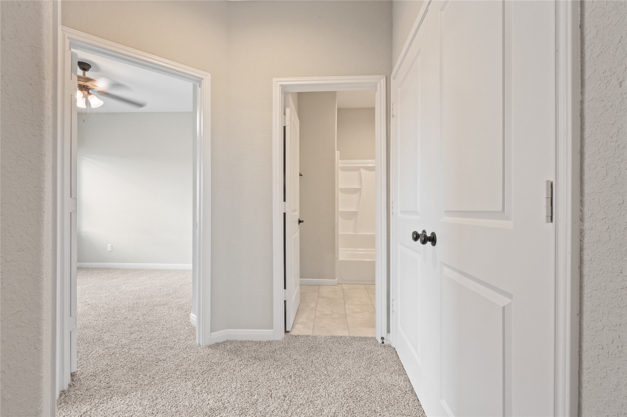 14010 Longstreet Road, Unit 2 Willis, TX 77318 - Photo 12 of 29 This photo shows a hallway with carpeted flooring leading to a bedroom with a ceiling fan and a bathroom. The walls are painted a neutral color, and there are white doors with dark knobs. The bathroom features a bathtub with built-in shelves.