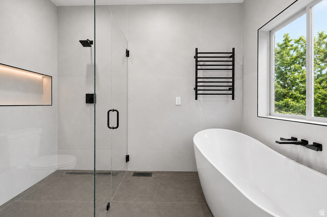 1429 Chateau Ridge Way Sandy, UT 84092 - Photo 10 of 22 Contemporary bathroom featuring a freestanding soaking tub, a spacious glass-enclosed shower with matte black fixtures, a wall-mounted towel warmer, a window with exterior foliage, and light-toned wall and floor tile