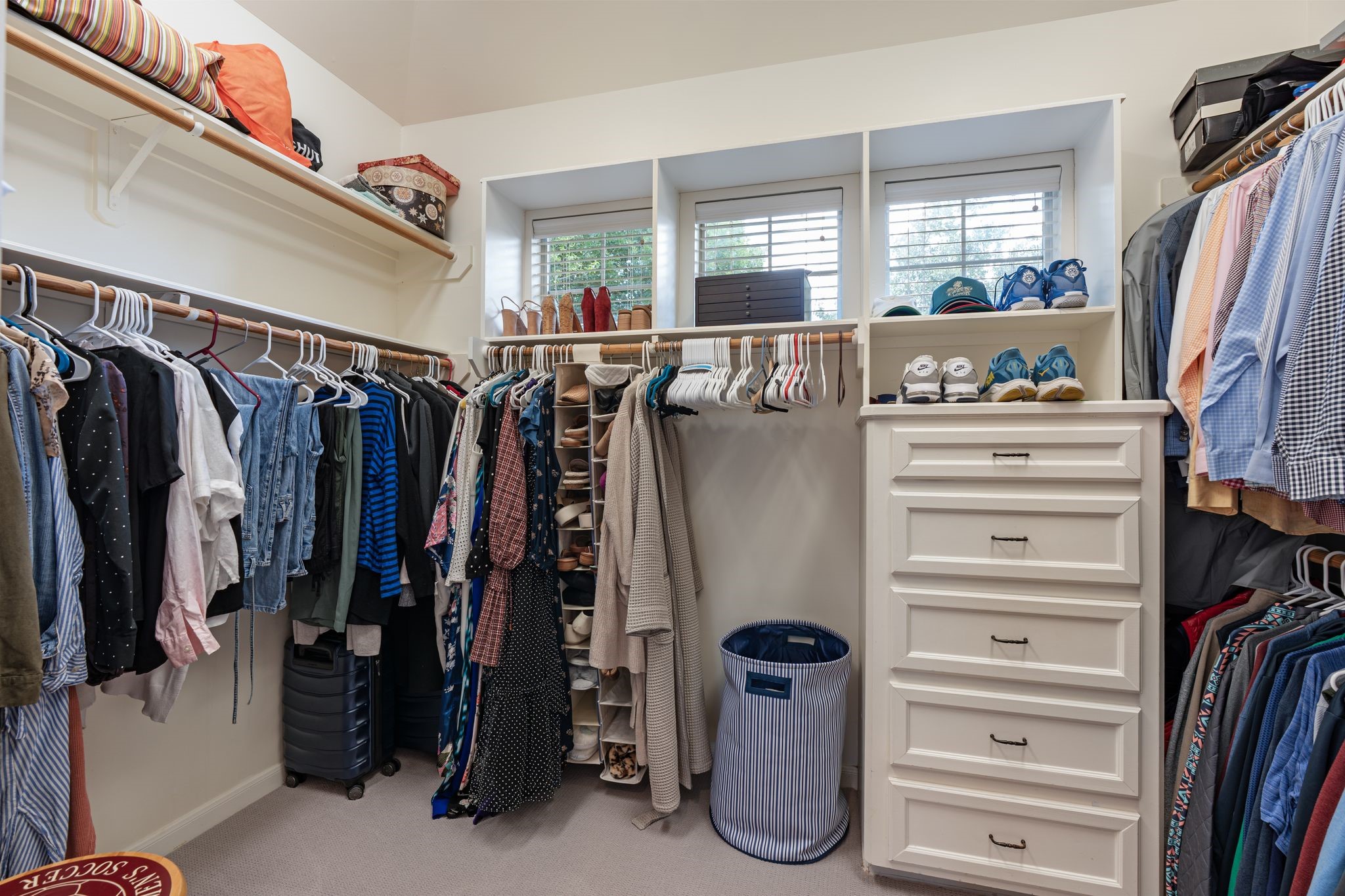 3822 Merrick Street Houston, TX 77025 - Photo 21 of 34 The primary walk-in closet offers extensive storage with dual hanging rods on multiple walls, upper shelving for bins and boxes, a built-in dresser with multiple drawers, additional cube-style shelving, and a natural light window with blinds. The carpeted space is well-proportioned for a shared closet.