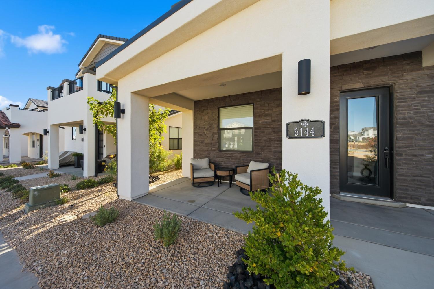 6144 Red Sand Lane St. George, UT 84790 - Photo 3 of 19 Step onto the covered porch and settle into the cushioned seating that flanks the front door — a perfect spot for morning coffee. Dark stacked tile, sleek black hardware, and the home's address plaque add refined character, while desert-adapted shrubs soften the entry with natural texture.