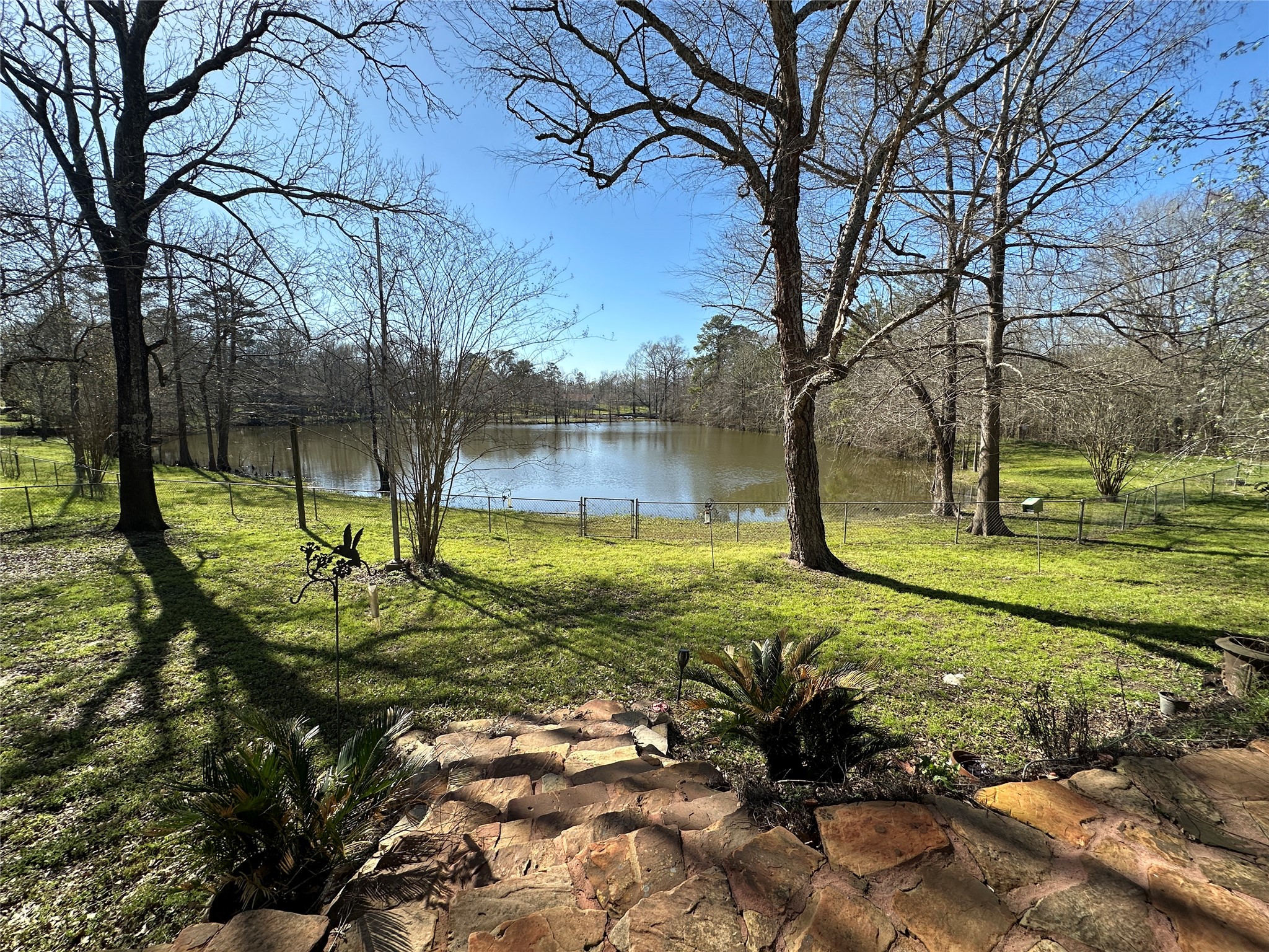 35 Susan Drive Cleveland, TX 77327 - Photo 30 of 33 This serene backyard features a picturesque view of a pond, surrounded by mature trees and lush grass. Stone steps lead down to the fenced area, offering a peaceful, natural setting perfect for relaxation and enjoying the outdoors.