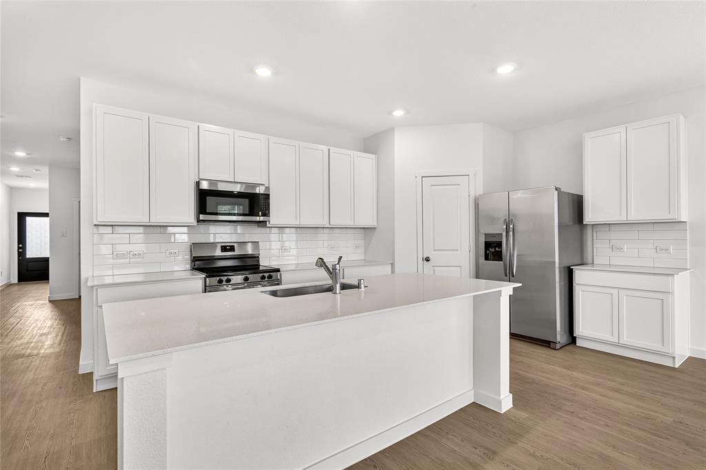 656 Waymaker Way Princeton, TX 75407 - Photo 5 of 40 Modern kitchen featuring an expansive center island with an undermount sink, white cabinetry, stainless steel appliances, a subway tile backsplash, and wood-finish flooring