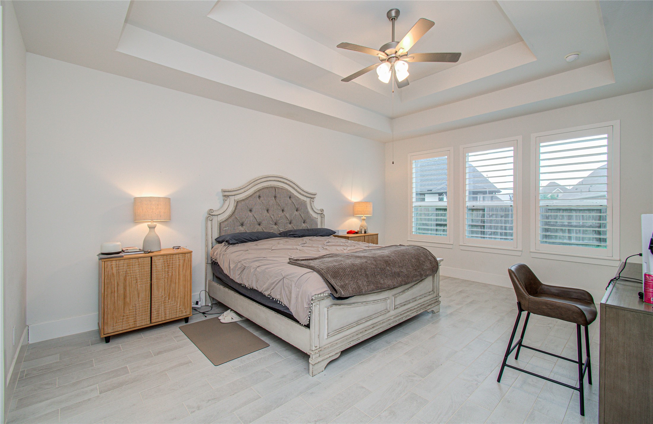 4211 Limestone Terrace Manvel, TX 77578 - Photo 16 of 33 Spacious bedroom with a tray ceiling and ceiling fan, featuring large windows with shutters for natural light. Includes a tufted bed, modern side tables with lamps, and a small desk with a chair.