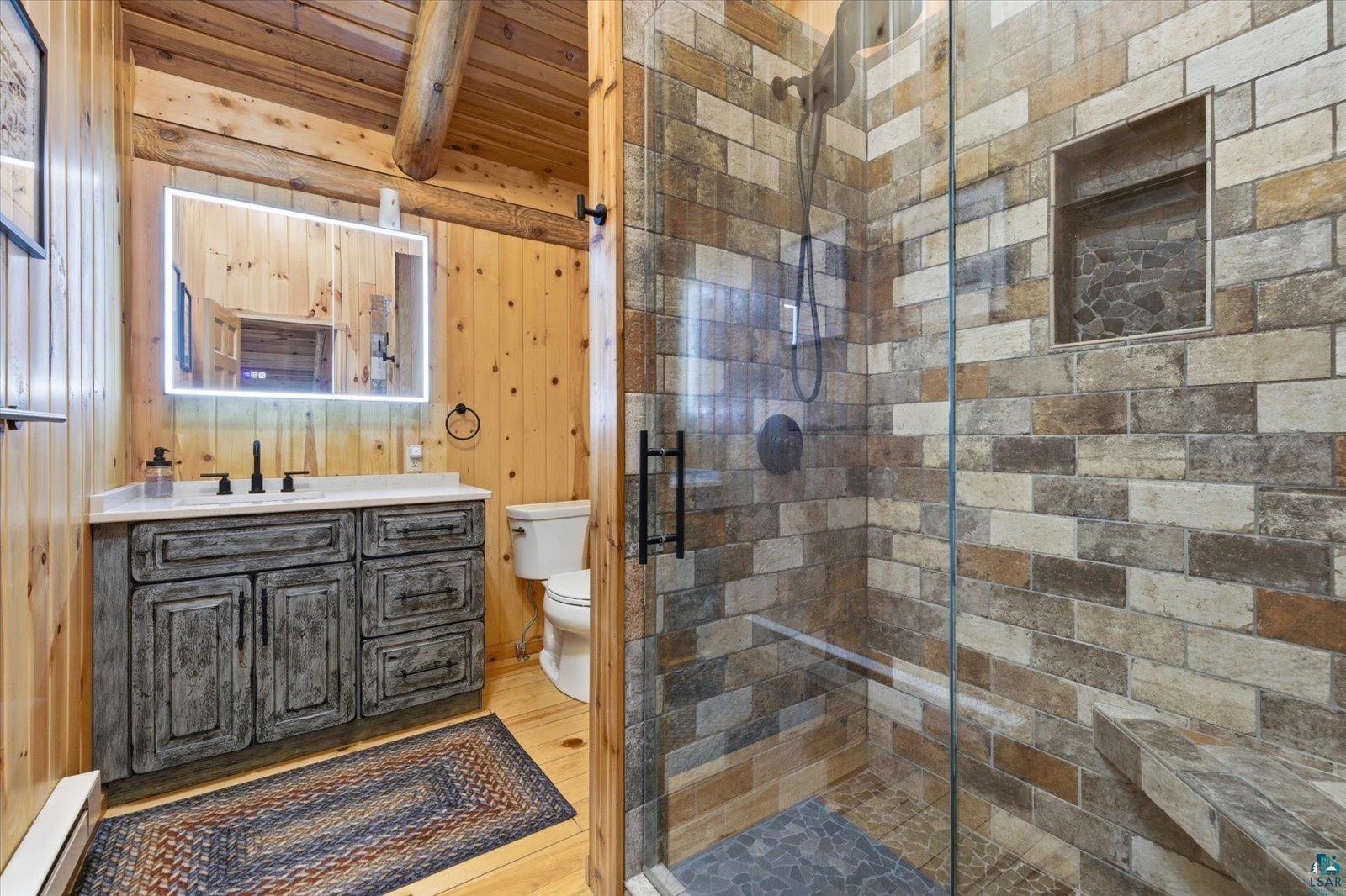 7185 Giants Ridge Road Embarrass, MN 55732 - Photo 31 of 47 Bathroom featuring tongue and groove wood paneling, a custom distressed wood vanity with a white countertop, an illuminated mirror, and a glass-enclosed shower with stacked stone tile, a built-in bench, and a recessed niche