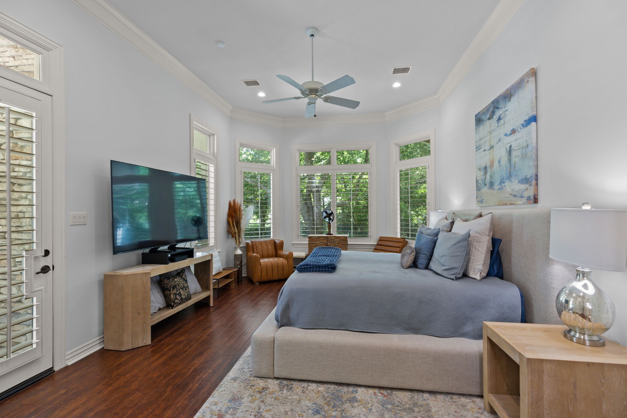 39 Maymont Way Spring, TX 77382 - Photo 17 of 50 This spacious bedroom features large windows with plantation shutters, offering plenty of natural light. It includes a cozy seating area, modern ceiling fan, and a mounted TV. The room is elegantly styled with hardwood floors and neutral tones, creating a serene atmosphere.
