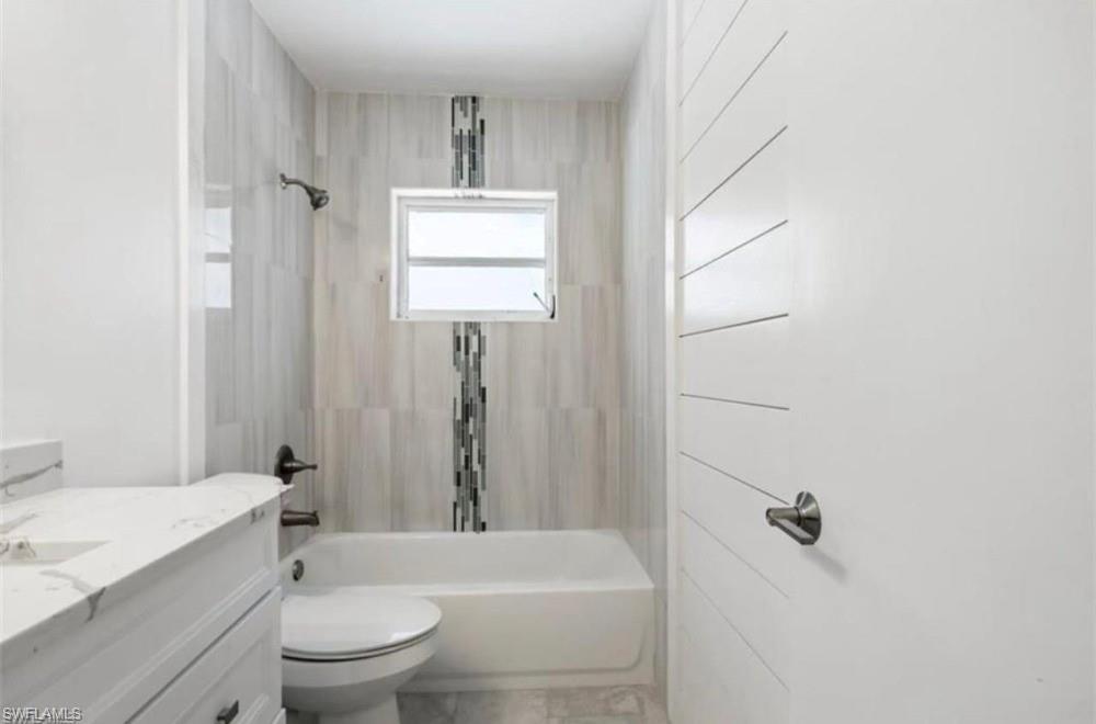 1948 46th Street Southwest Naples, FL 34116 - Photo 11 of 14 Bathroom featuring a built-in tub with a shower fixture, light-toned vertical wall tiling with a mosaic accent, a small window, a white vanity with a marble-finish countertop, and a toilet