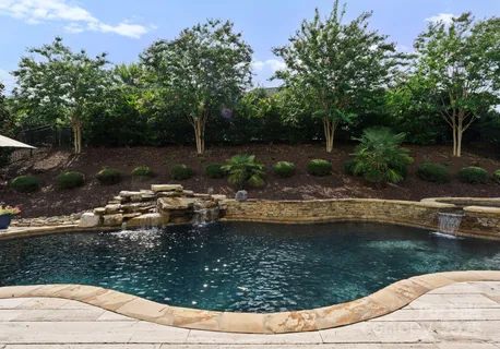 a view of a swimming pool with lawn chairs plants and large trees