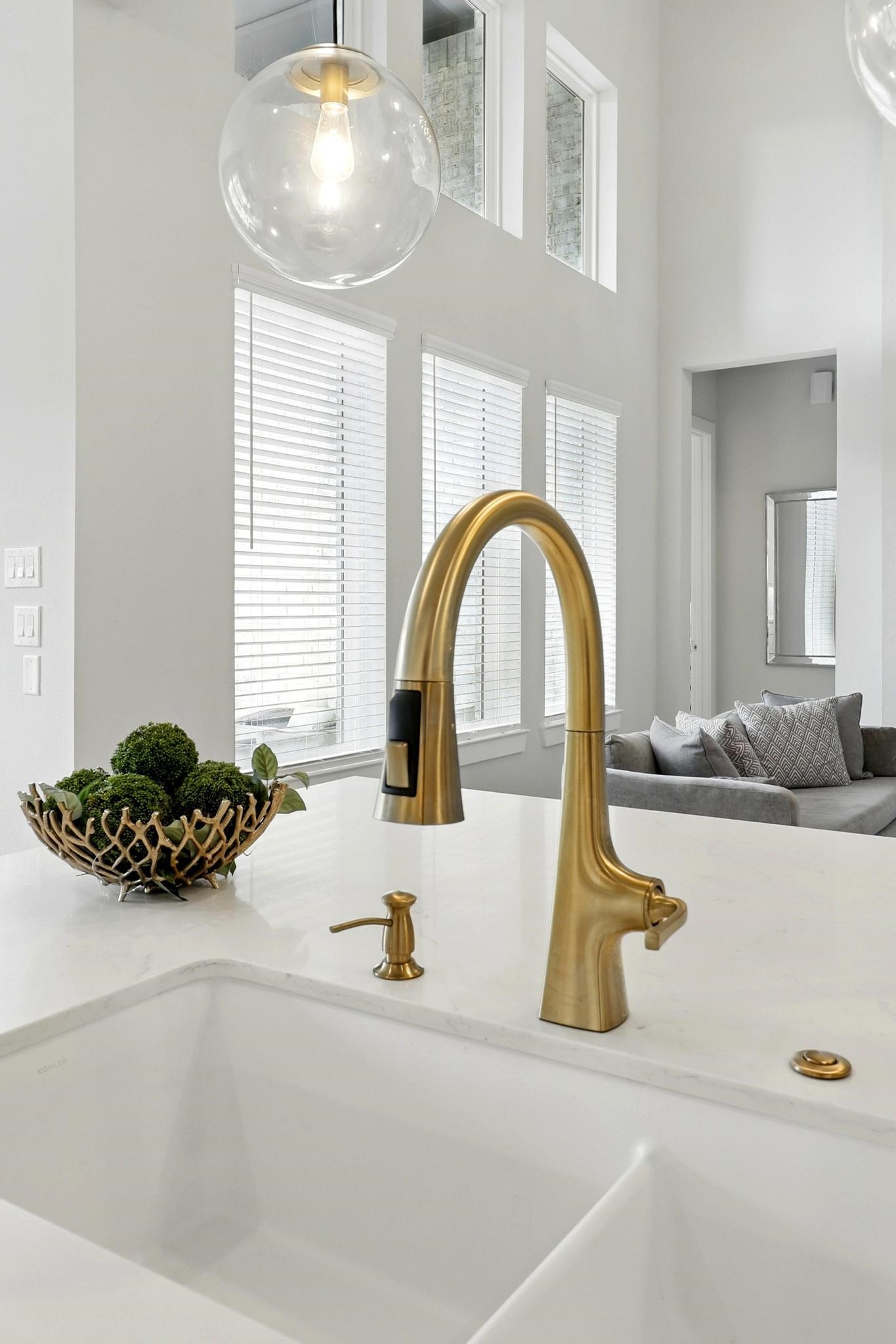 26923 Arethusa Court Spring, TX 77389 - Photo 25 of 44 This modern kitchen features a sleek gold faucet on a white countertop, with ample natural light from large windows and a cozy living space visible in the background.