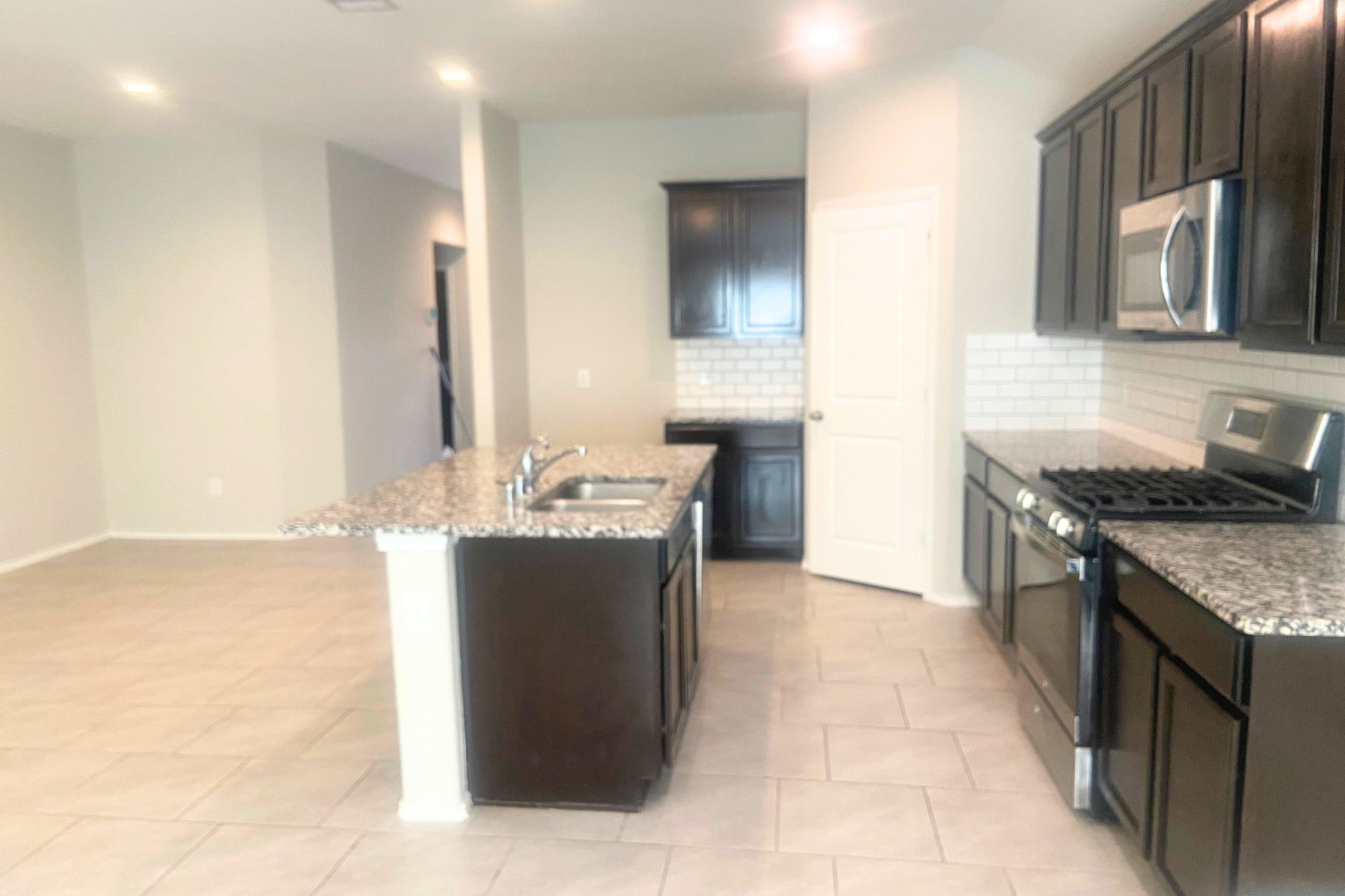 8310 Rose Petals Lane Rosharon, TX 77583 - Photo 5 of 43 Open view of the modern kitchen from the living area, featuring a large granite island, dark wood cabinetry, stainless steel appliances, and elegant subway tile backsplash. The open-concept layout with recessed lighting and tile flooring creates a bright, spacious area perfect for everyday living and entertaining.