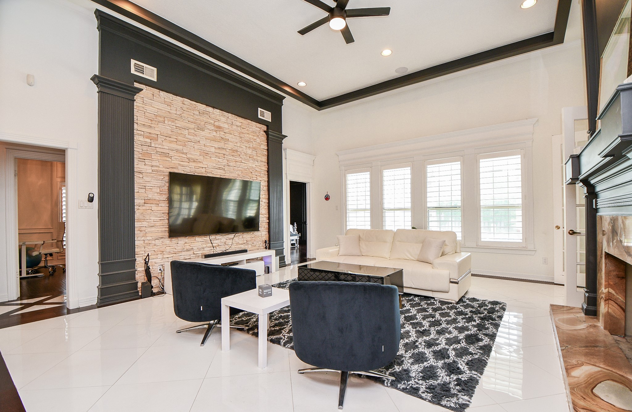 263 Wood Loop Street Houston, TX 77015 - Photo 21 of 45 This spacious living room features high ceilings with modern recessed lighting and a ceiling fan. It includes a striking stone accent wall with a mounted flat-screen TV, a cozy fireplace, and large windows that allow natural light to fill the space. The room is elegantly furnished with a white leather sofa and modern chairs on a stylish rug, creating a welcoming atmosphere.
