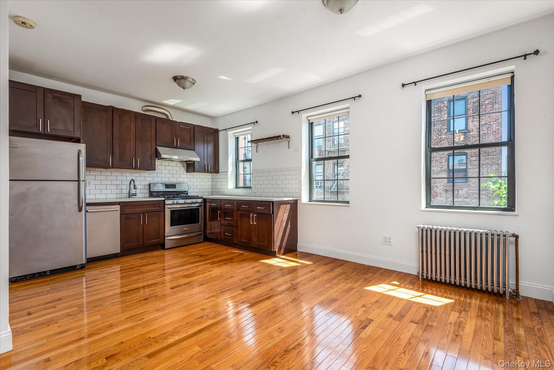 26-19 14th Place Queens, NY 11102 - Photo 21 of 33 Kitchen featuring appliances with stainless steel finishes, radiator, light wood-style floors, backsplash, and dark brown cabinets