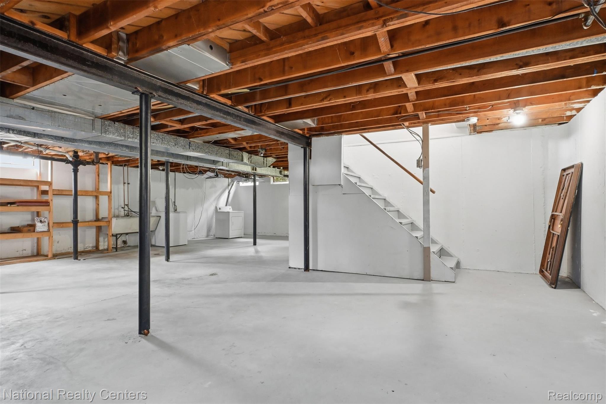 17159 Rougeway Street Livonia, MI 48152 - Photo 29 of 36 Spacious unfinished basement featuring exposed wood joists and structural beams, painted concrete flooring, a white staircase with wood handrail, and built-in wooden shelving