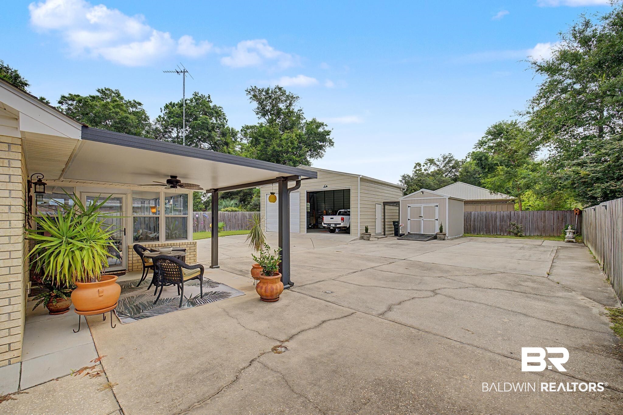 2808 Pine Ridge Drive Lillian, AL 36549 - Photo 29 of 47 Expansive concrete driveway leading to a covered patio with a ceiling fan, a detached metal-sided garage, and a separate storage shed, all bordered by a wood fence