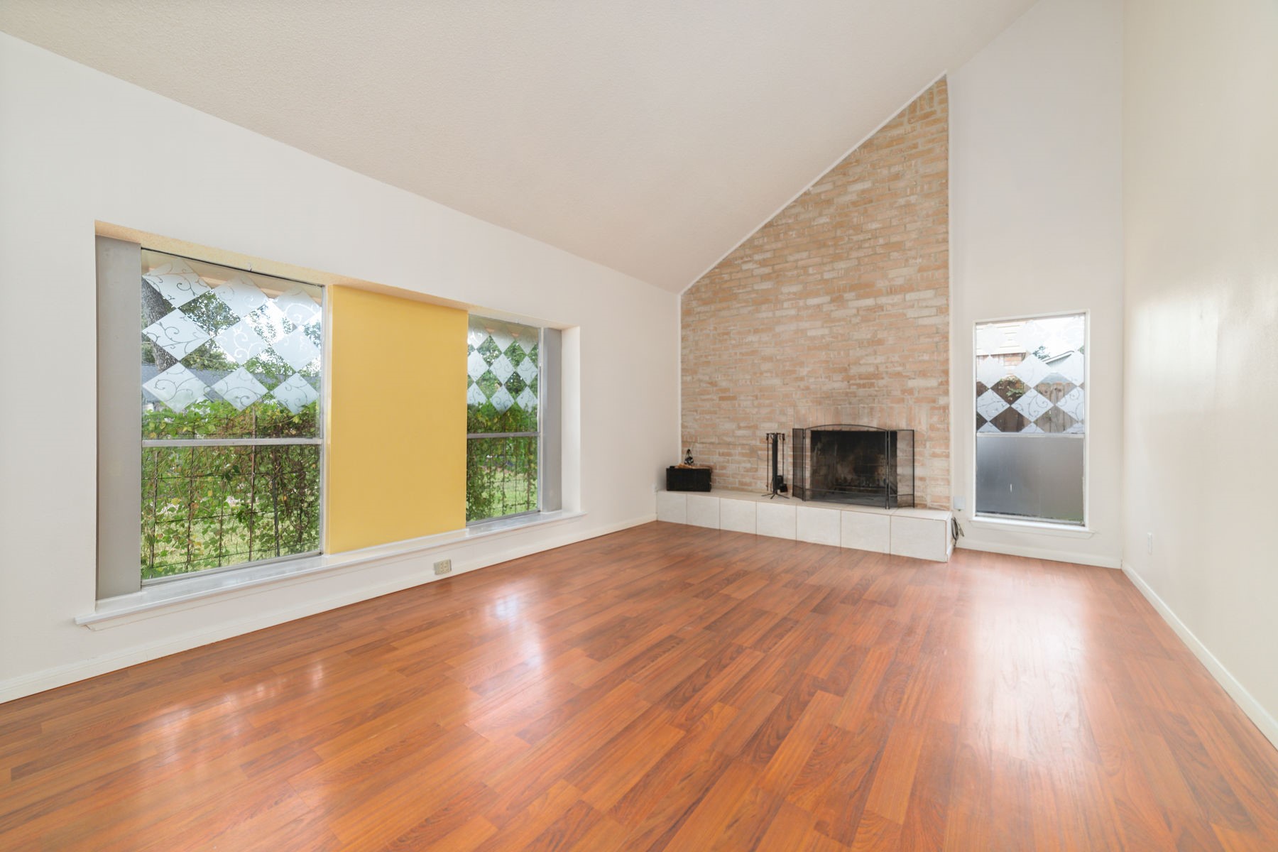 25402 Mill Pond Lane Spring, TX 77373 - Photo 9 of 24 This spacious living room features high ceilings, a brick accent wall with a fireplace, large decorative windows for natural light, and warm wood flooring, creating an inviting and cozy atmosphere.
