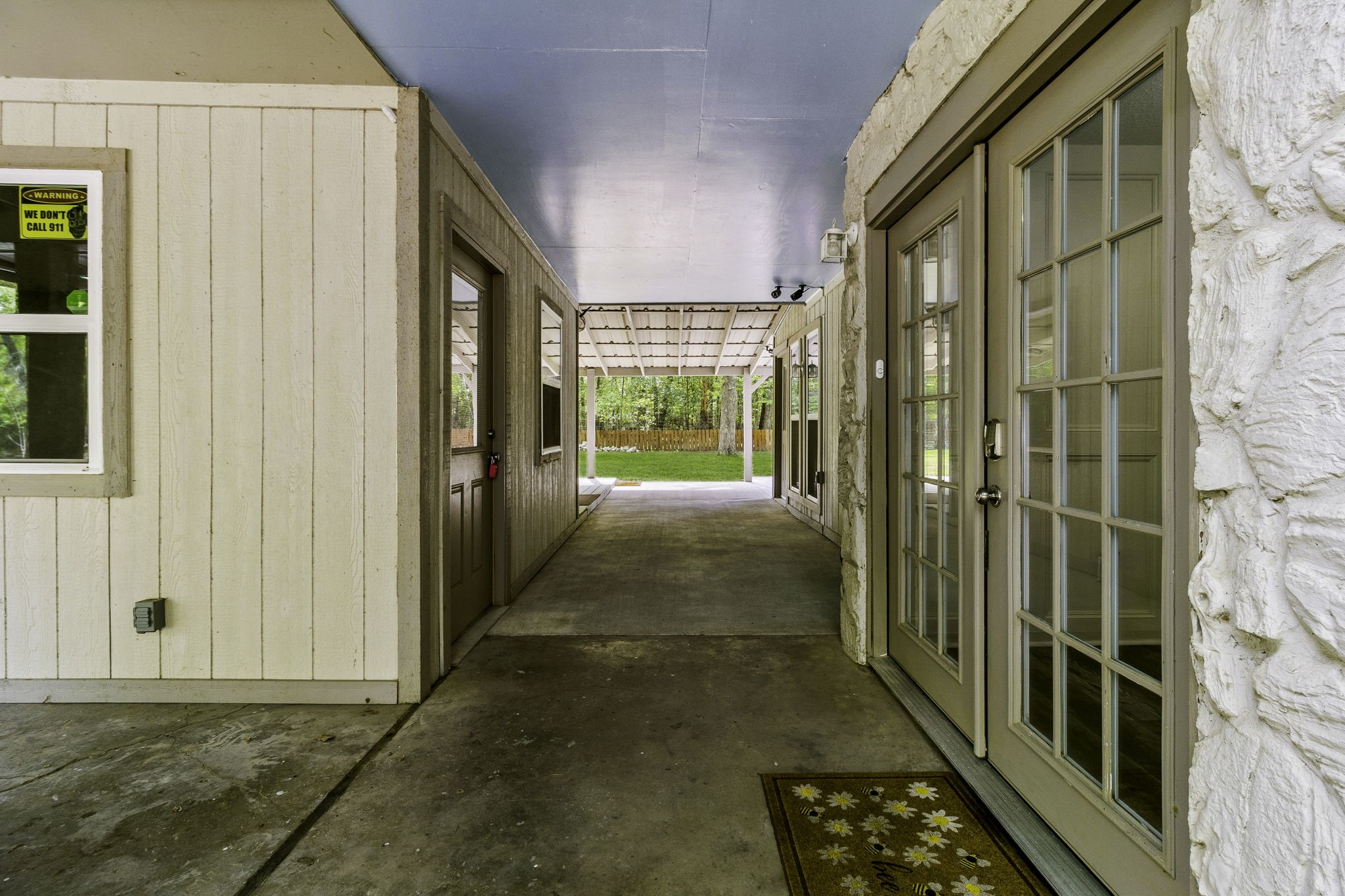 21 Red Oak Circle Point Blank, TX 77364 - Photo 18 of 32 This photo shows a covered outdoor space with a concrete floor, leading to a backyard. It features beige siding, large glass French doors, and a ceiling with a soft blue tone. The area provides a sheltered transition between indoor and outdoor living spaces.