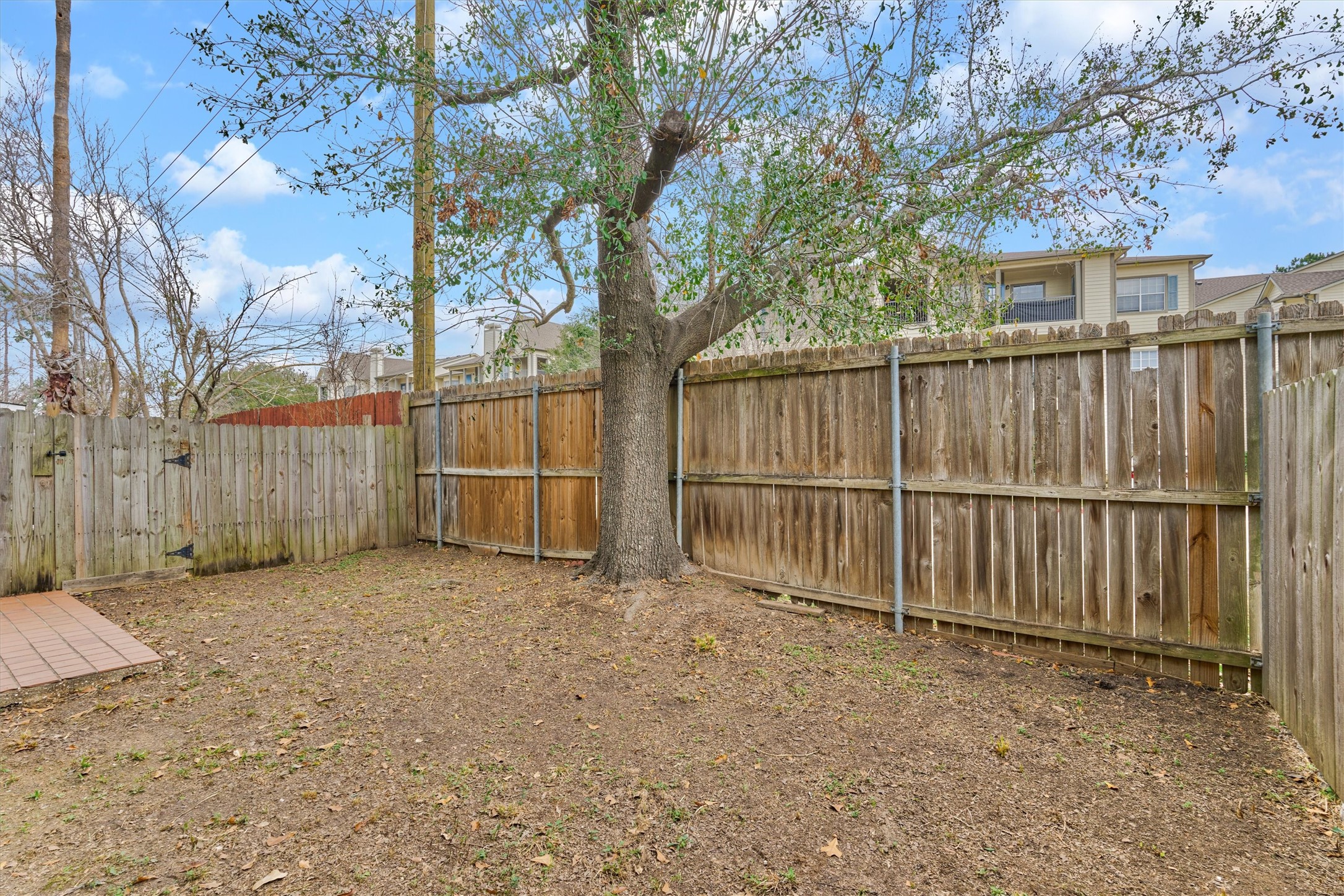 14420 Walters Road, Unit 26 Houston, TX 77014 - Photo 40 of 45 This backyard features a wooden fence for privacy, a central mature tree providing shade, and a small tiled patio area, perfect for outdoor activities.