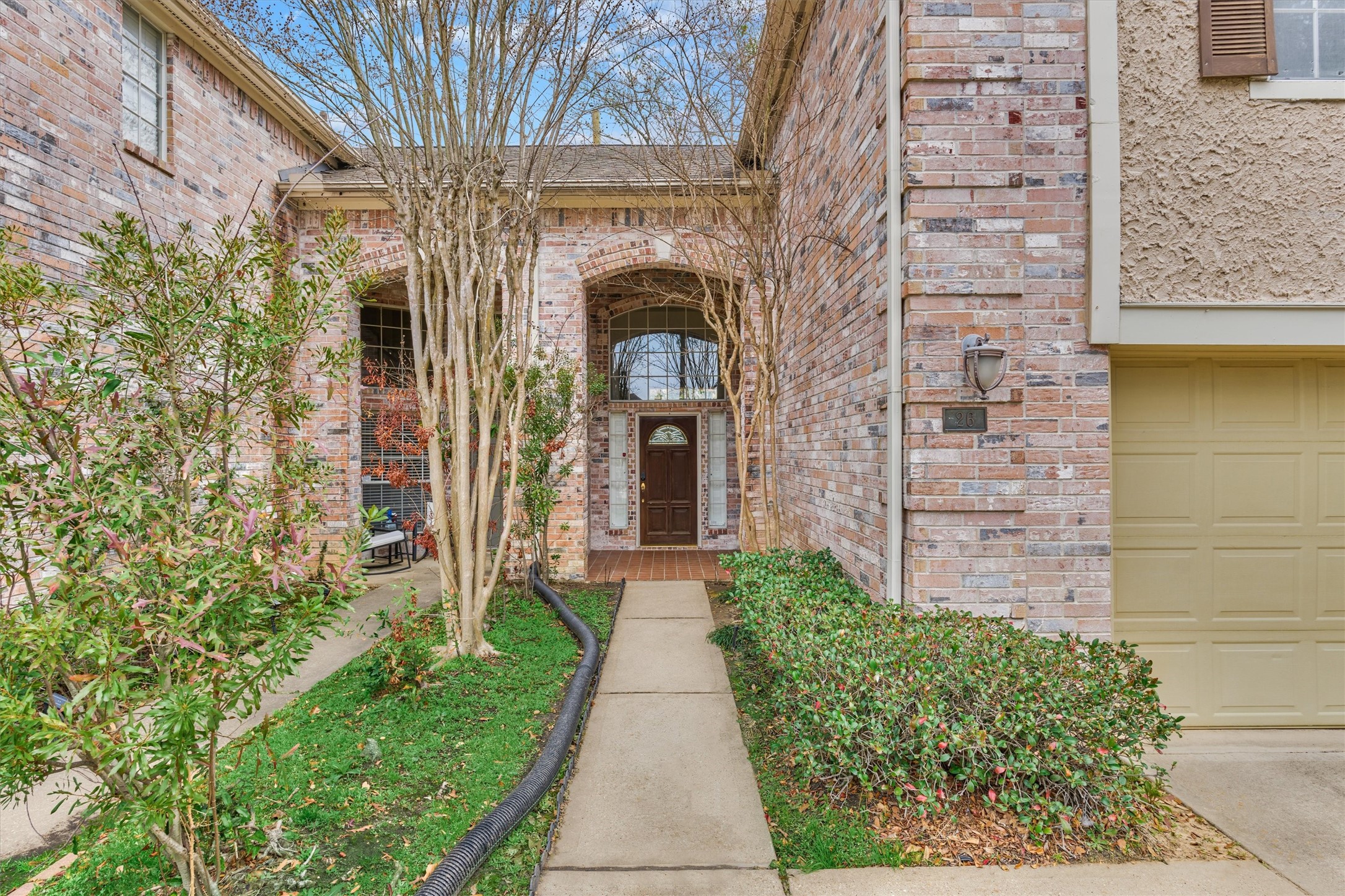 14420 Walters Road, Unit 26 Houston, TX 77014 - Photo 4 of 45 This photo shows a welcoming brick home exterior with a charming front entryway. The path is lined with shrubs and trees, leading to a door with a glass insert. The garage is visible on the right side.
