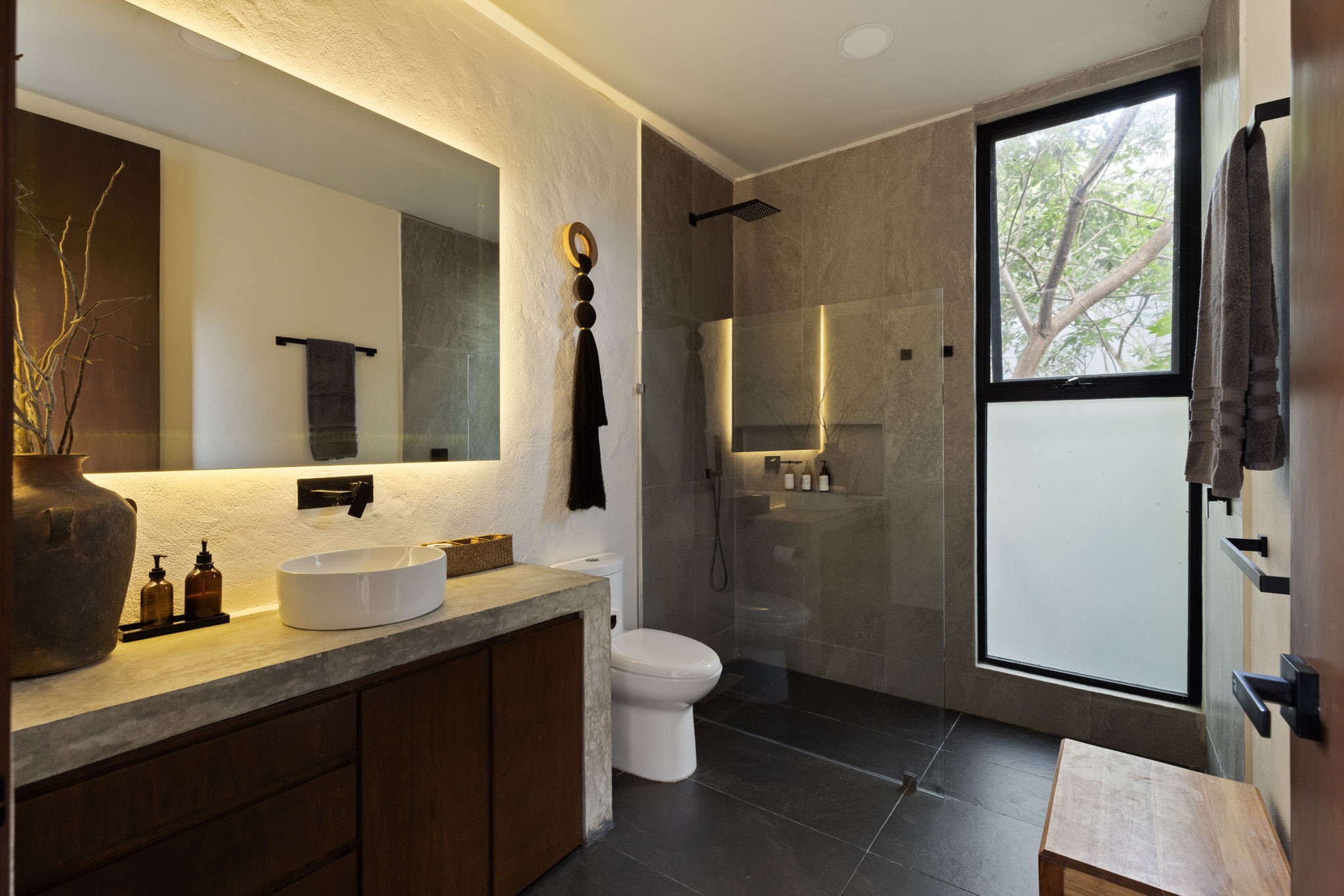 6 Country Club, Unit 4 Nome, TX 77629 - Photo 25 of 45 This exquisitely appointed secondary bathroom showcases a resort-style design that perfectly matches the home's signature contemporary aesthetic and organic material palette. The luxurious space features a spacious wet room wrapped in large-format stone tiles with a frameless glass entry, while a large vertical window draws in natural daylight and frames views of the tropical treetops. The gorgeous vanity completes this bathroom with a custom-cast concrete countertop and modern white vessel sink.
