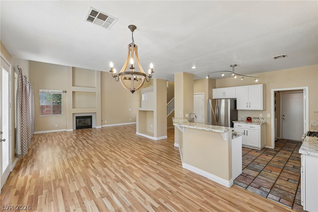1727 Millstream Way Henderson, NV 89074 - Photo 15 of 46 A striking chandelier hangs over a spacious room with light hardwood flooring, complemented by a kitchen featuring a peninsula and tiled flooring.