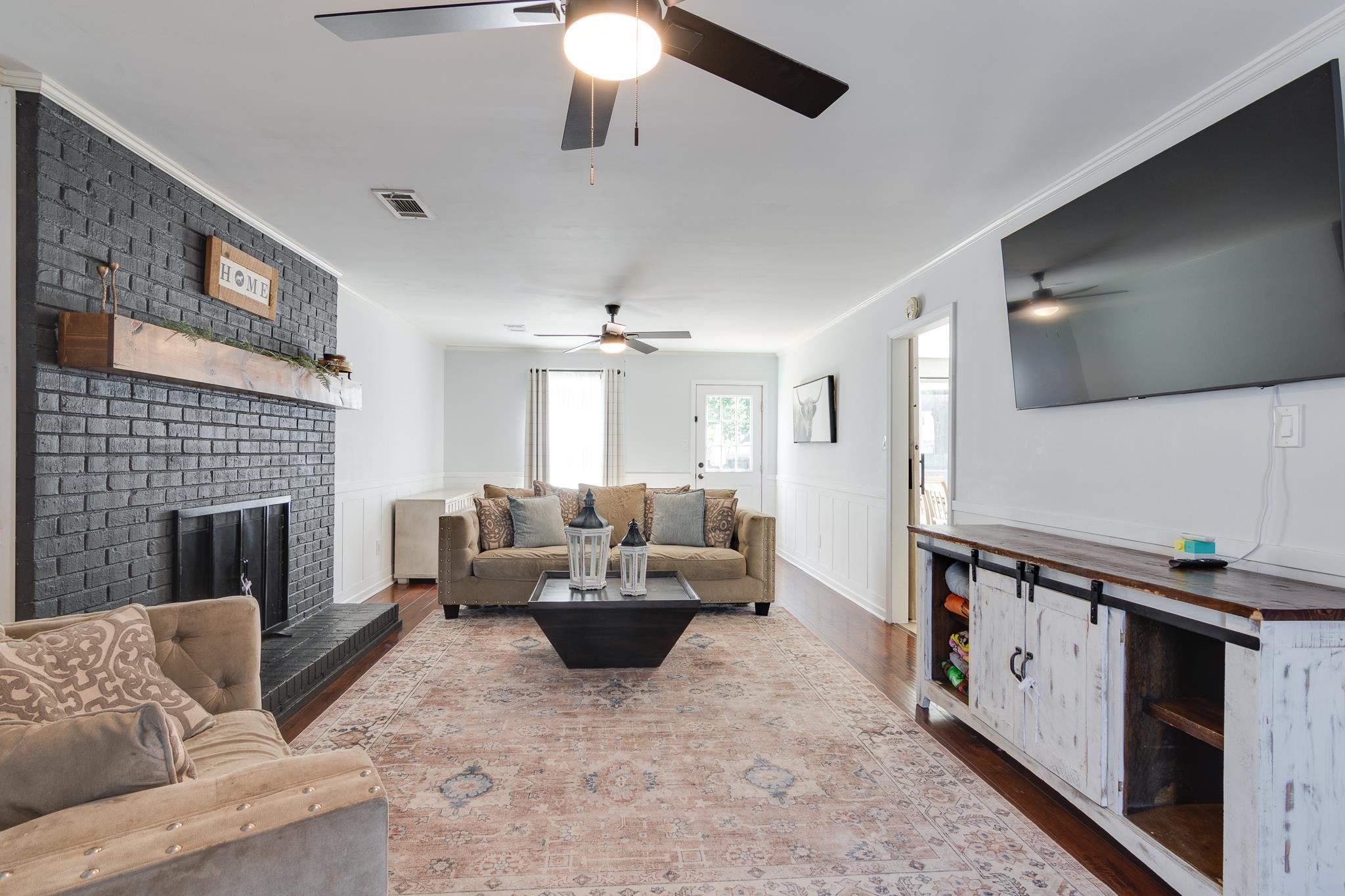 Spacious room featuring a painted brick fireplace with wood mantel, a built-in media console with sliding barn doors, two ceiling fans, and wood-finish flooring