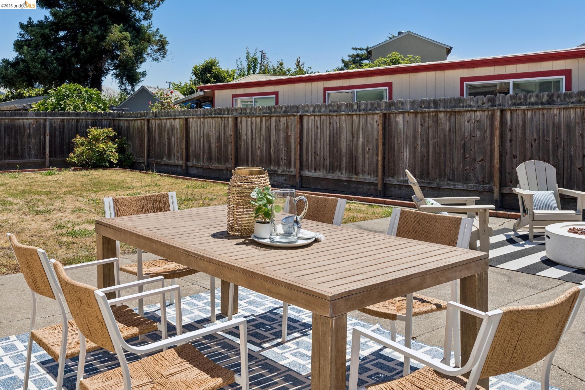 15162 Inverness Street San Leandro, CA 94579 - Photo 19 of 25 Concrete patio featuring a slatted outdoor dining table, a patterned area rug, and a separate seating area with Adirondack chairs and a fire pit