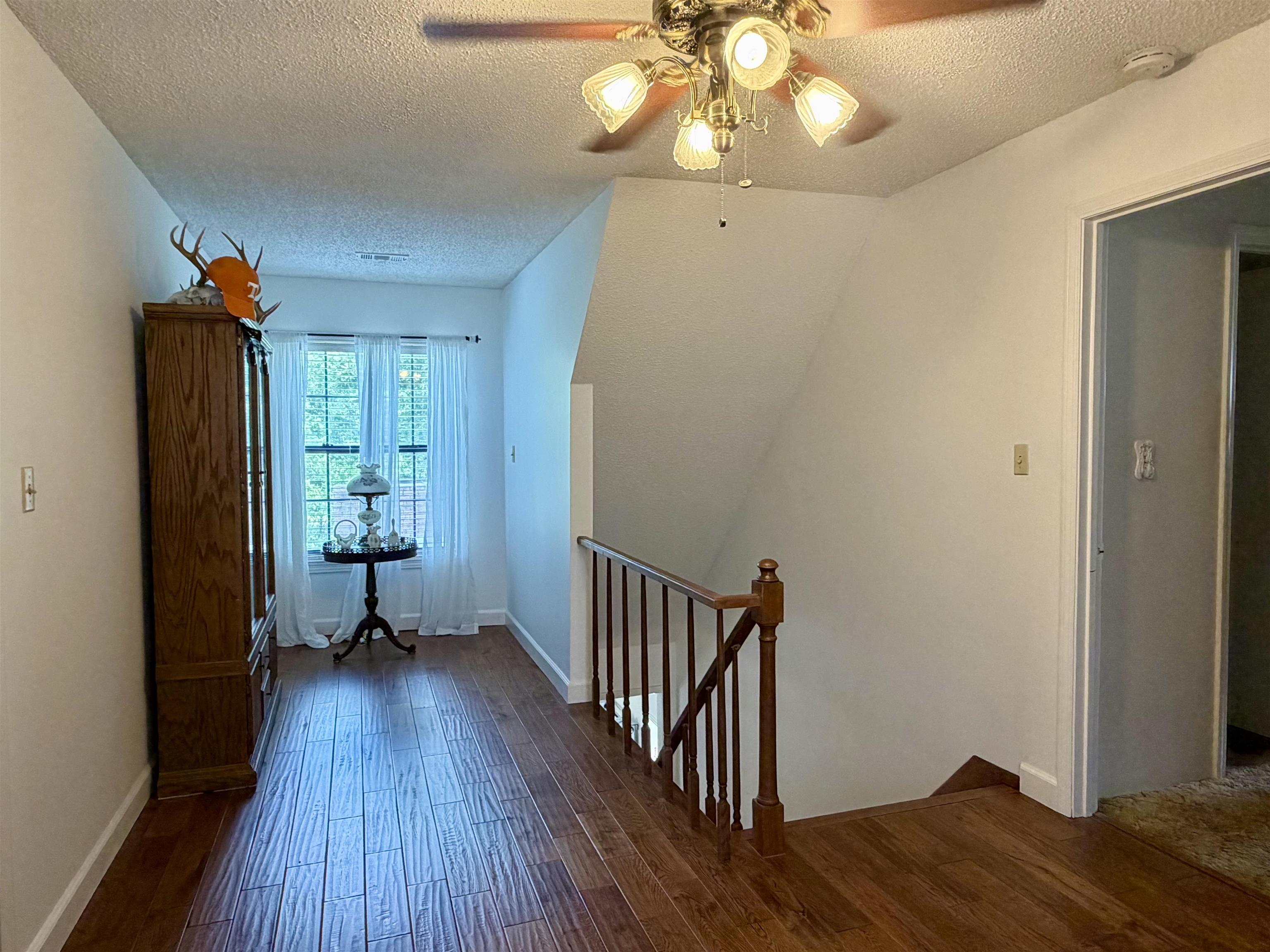 142 Treece Street Selmer, TN 38375 - Photo 15 of 36 Hallway featuring wood-finish flooring, a window with sheer curtains, a wooden staircase with railing, and a ceiling fan with light fixture