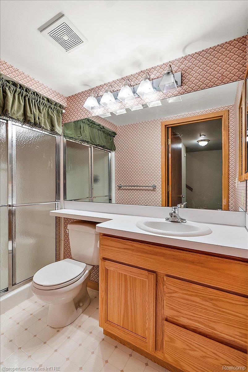 6175 Tripp Road Holly, MI 48442 - Photo 34 of 63 Bathroom featuring a wood-finish vanity with a white countertop and integrated sink, a large wall-mounted mirror, a shower stall with clear glass doors, and patterned wallpaper
