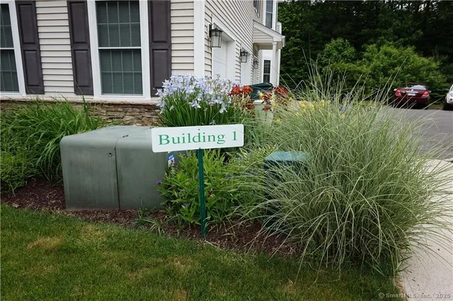 a sign that is sitting in front of a house with plants and trees