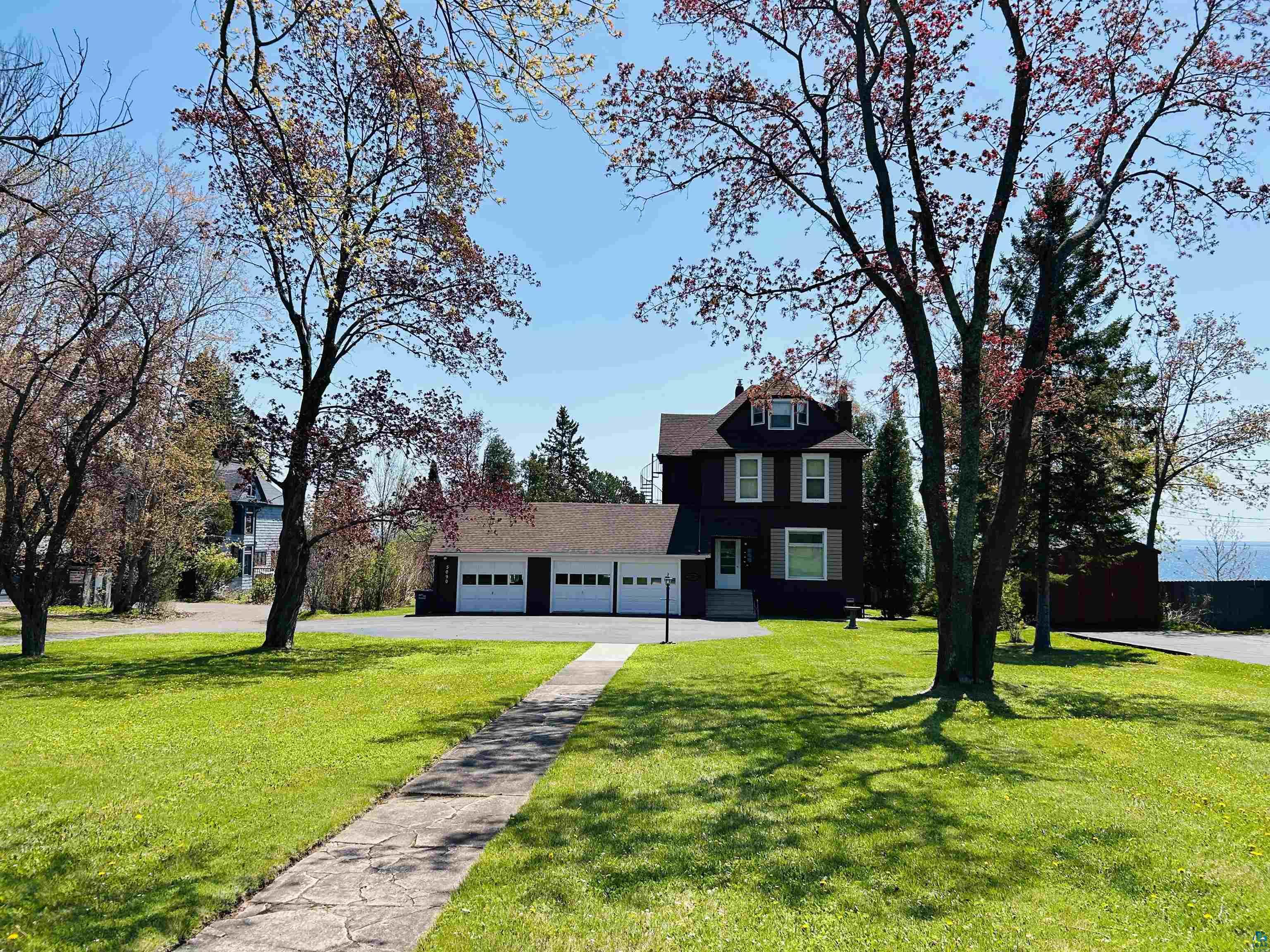 5090 London Road, Unit 1 Duluth, MN 55804 - Photo 2 of 54 Exterior featuring a dark-colored facade with white trim, a multi-car garage, established front lawn, mature trees, and a concrete pathway