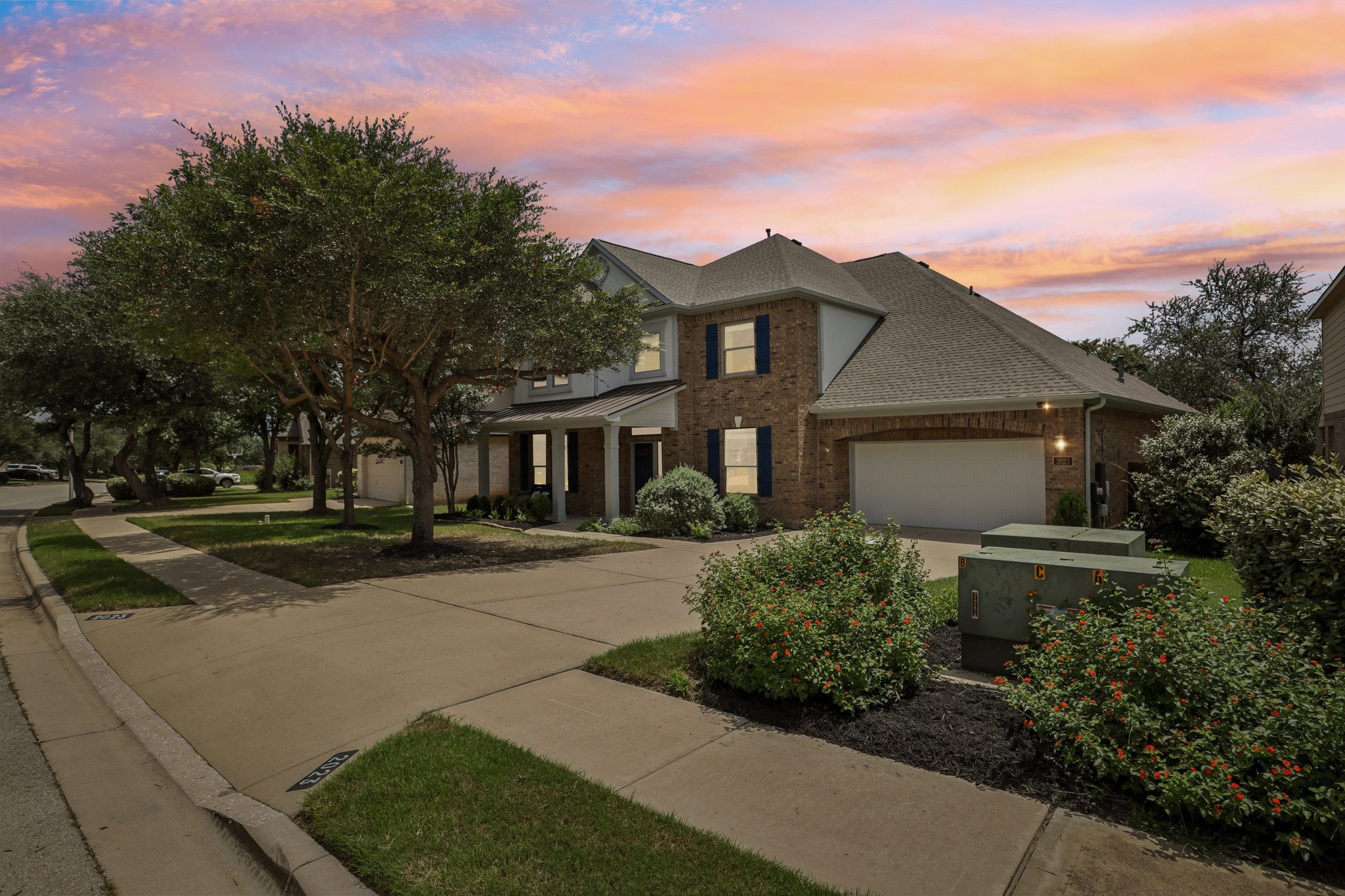 2023 Nelson Ranch Loop Cedar Park, TX 78613 - Photo 39 of 40 A classic brick-front home with blue window shutters and a two-car garage sits under a vibrant twilight sky.