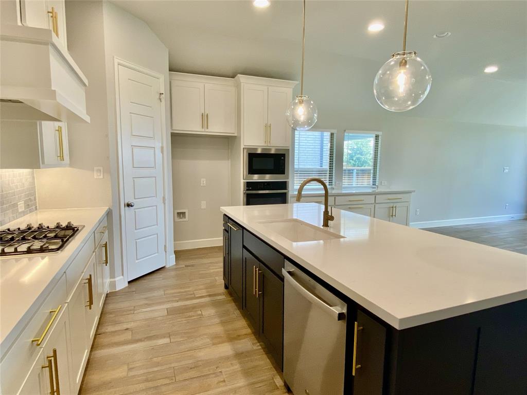 1937 Halkin Road Celina, TX 75009 - Photo 13 of 20 Contemporary kitchen featuring a large central island with an undermount sink, pendant lighting, built-in stainless steel appliances, white cabinetry with gold-tone hardware, and wood-look tile flooring