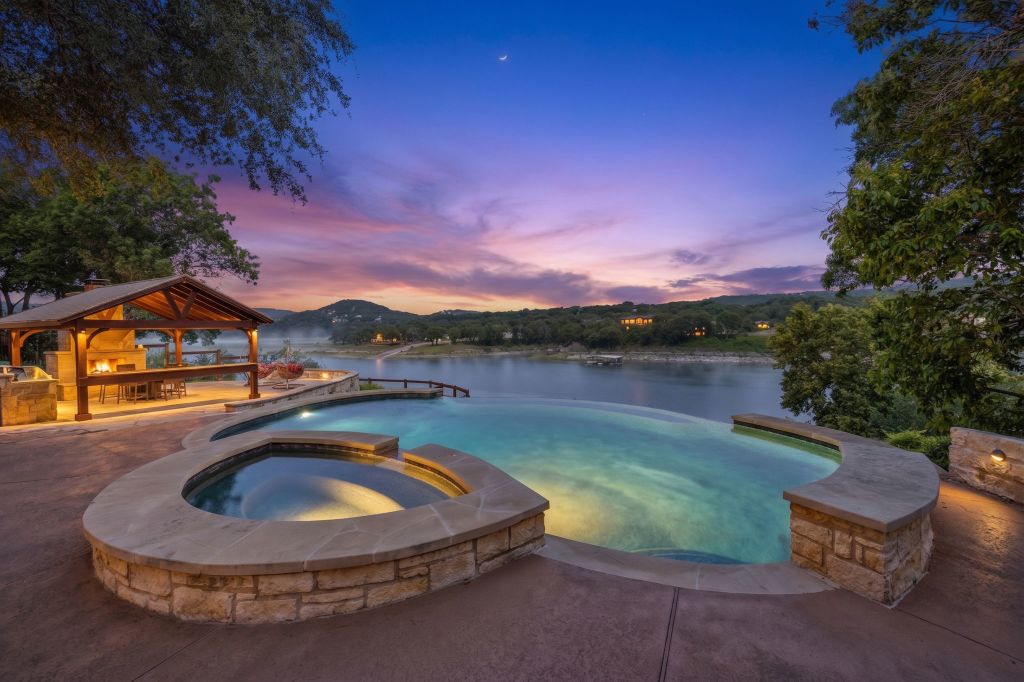 An exquisite infinity-edge pool and spa offer tranquil lake views at sunset, complemented by a covered outdoor kitchen and stone fireplace.