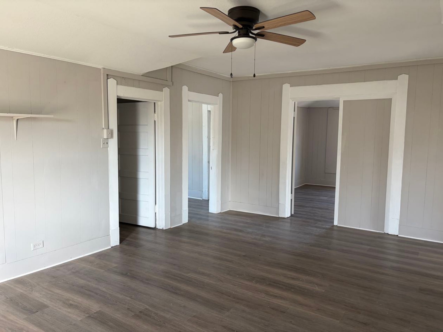 931 9th Avenue, Unit 2 Port Arthur, TX 77642 - Photo 1 of 9 Living Room - Spacious room with light gray paneled walls, dark wood flooring, and a ceiling fan. It features three doorways leading to adjacent rooms, providing an open flow layout.