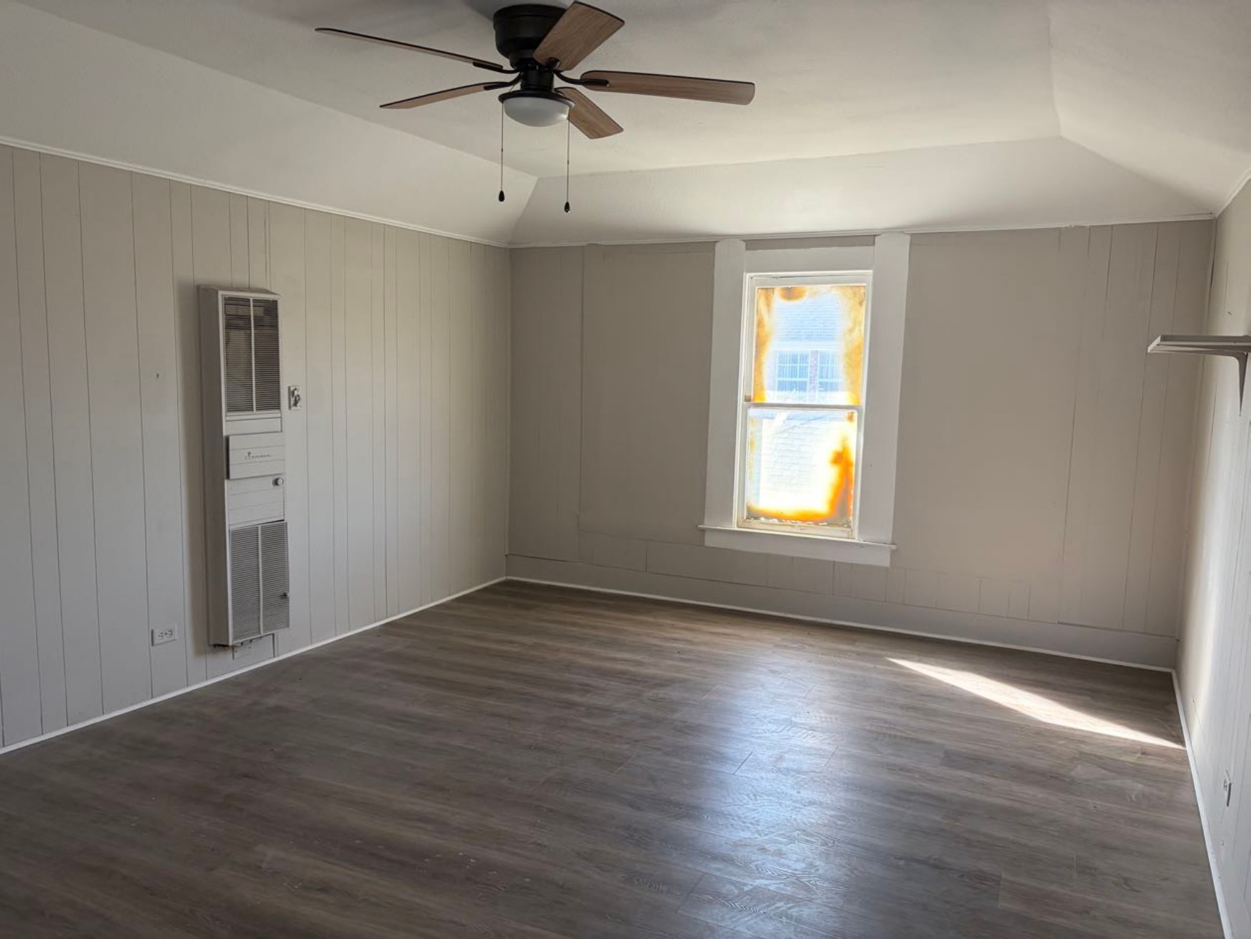 931 9th Avenue, Unit 2 Port Arthur, TX 77642 - Photo 2 of 9 This room features neutral-toned walls with vertical paneling, a ceiling fan, and a wall-mounted heater. A large window allows natural light to illuminate the space, highlighting the wood-style flooring.