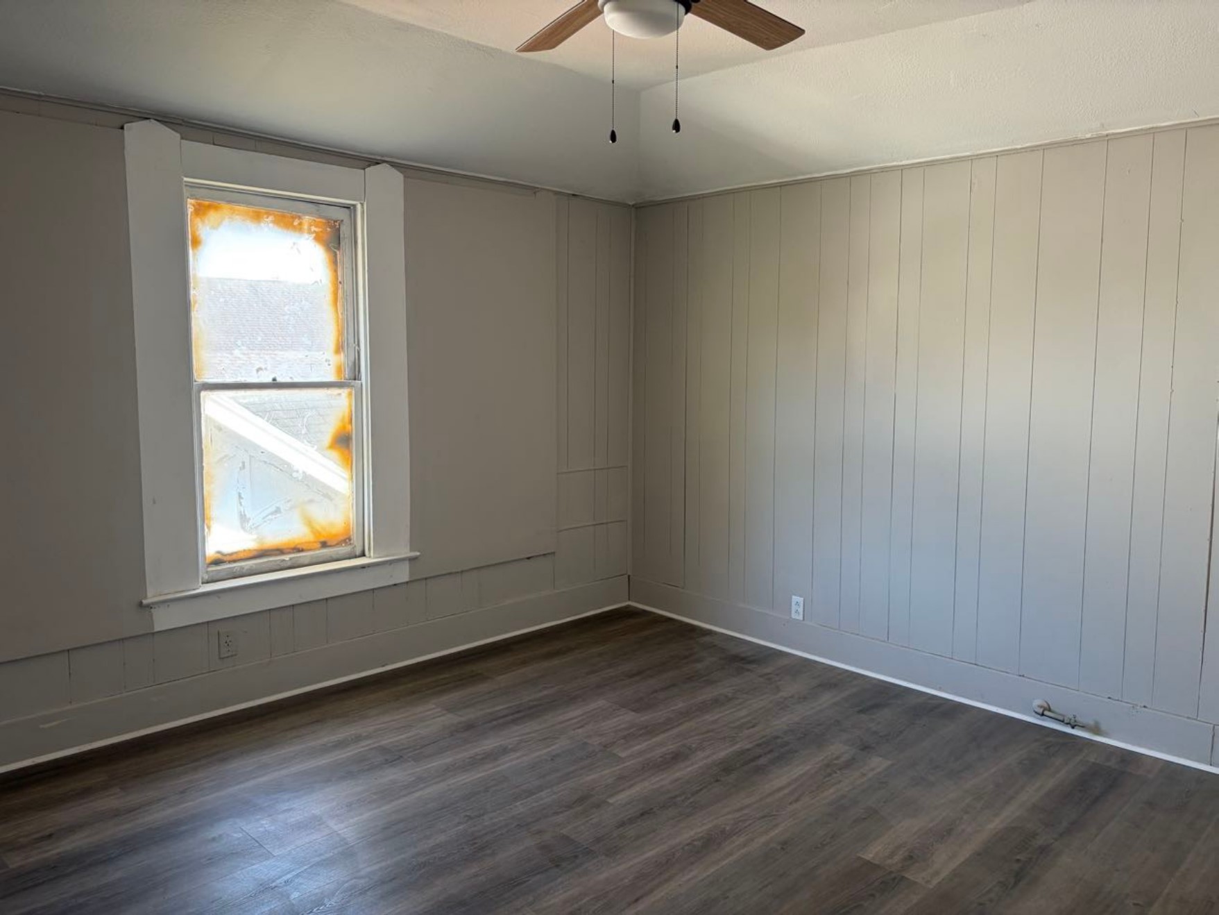 931 9th Avenue, Unit 2 Port Arthur, TX 77642 - Photo 3 of 9 This room features paneled walls painted in a neutral tone, a ceiling fan, and laminate wood flooring. The window allows natural light, but the frame shows signs of wear. Overall, it’s a simple, functional space with potential for customization.
