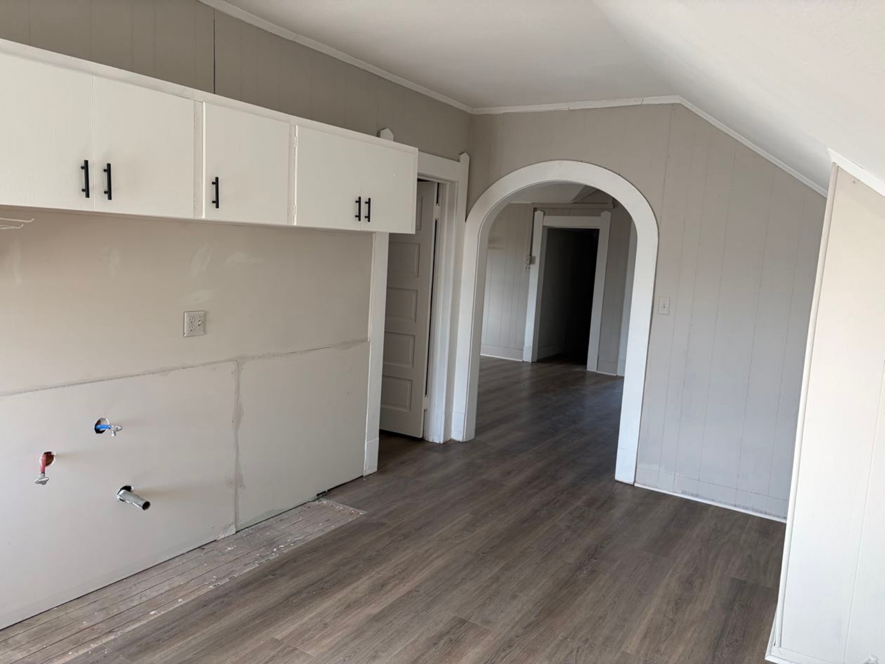 931 9th Avenue, Unit 2 Port Arthur, TX 77642 - Photo 8 of 9 Kitchen - This photo shows a room with laminated flooring. It features white upper cabinets with black handles and visible plumbing hookups, likely for appliances. An arched doorway leads to an adjoining space, adding architectural interest.