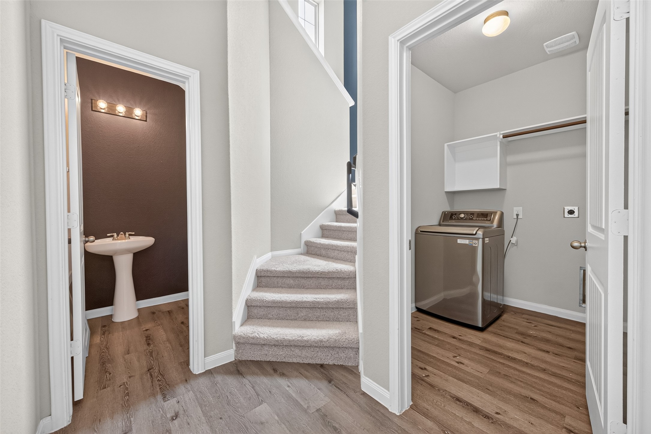 2526 Sturgeon Way Conroe, TX 77384 - Photo 15 of 49 This photo showcases a clean and modern hallway with light wood flooring. It features a half-bathroom with a pedestal sink on the left, a carpeted staircase leading upstairs, and a laundry room with a washing machine on the right. Perfect for convenient living.
