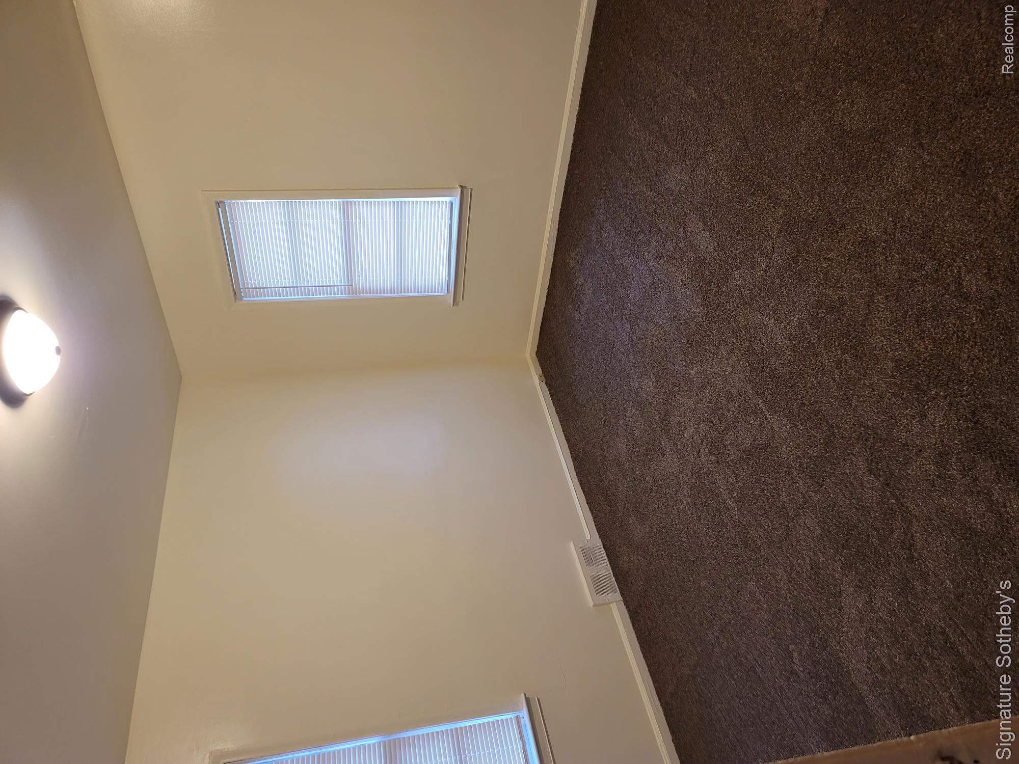 Neutral-toned walls, two windows with blinds, dark textured carpeting, and a ceiling-mounted light fixture
