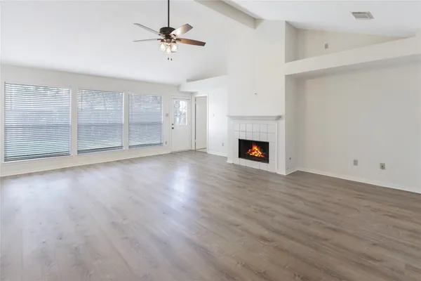 an empty room with wooden floor a fireplace a ceiling fan and a kitchen view