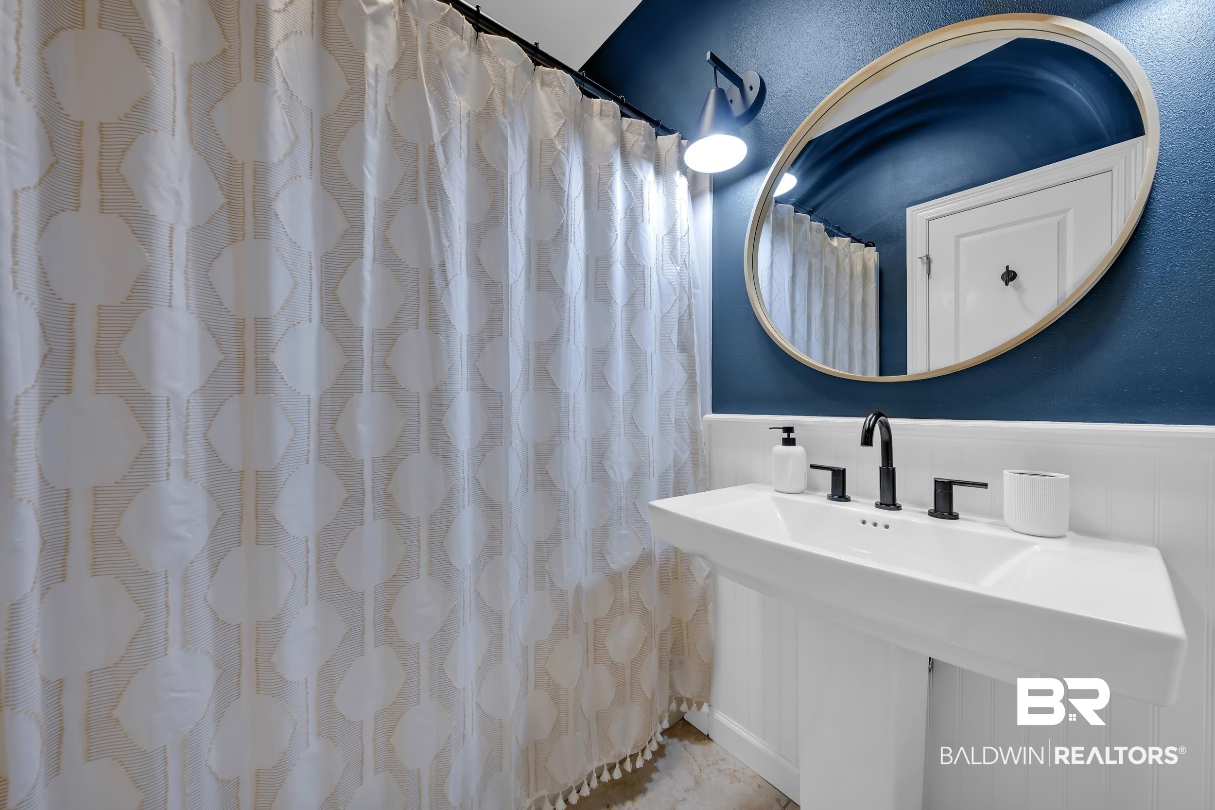 8137 Pine Run Daphne, AL 36527 - Photo 36 of 67 Pedestal sink with matte black faucet, oval mirror, wall-mounted light fixture, blue wall, and white wainscoting